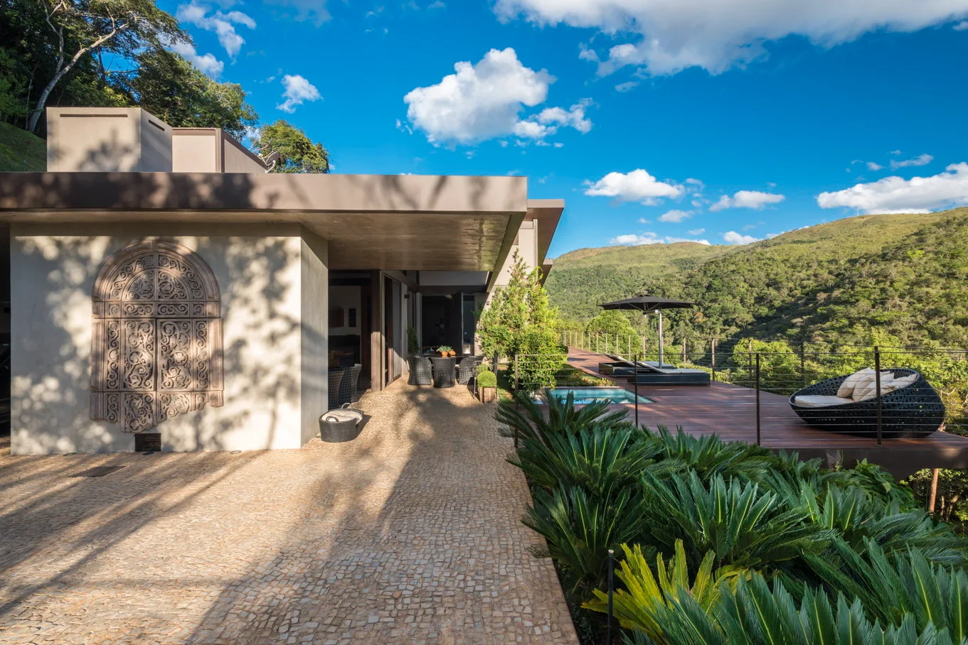 A modern outdoor terrace features a cobblestone path, a carved stone wall relief, and a wood deck with a swimming pool overlooking hills.