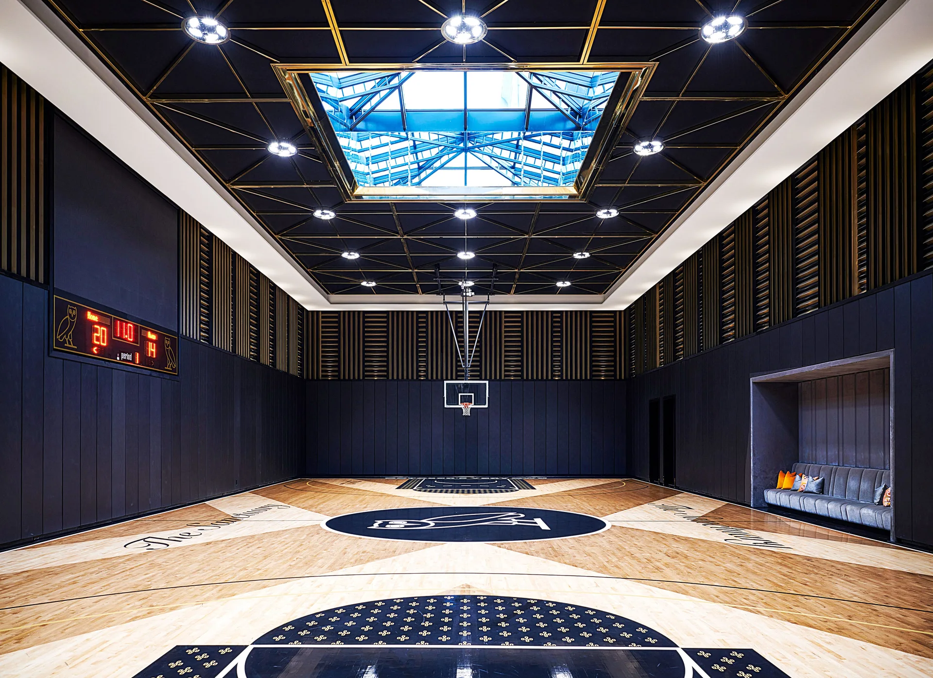 An indoor basketball court features a light wood floor, matte black paneled walls, a built-in seating alcove, and a black ceiling with gold geometric details.