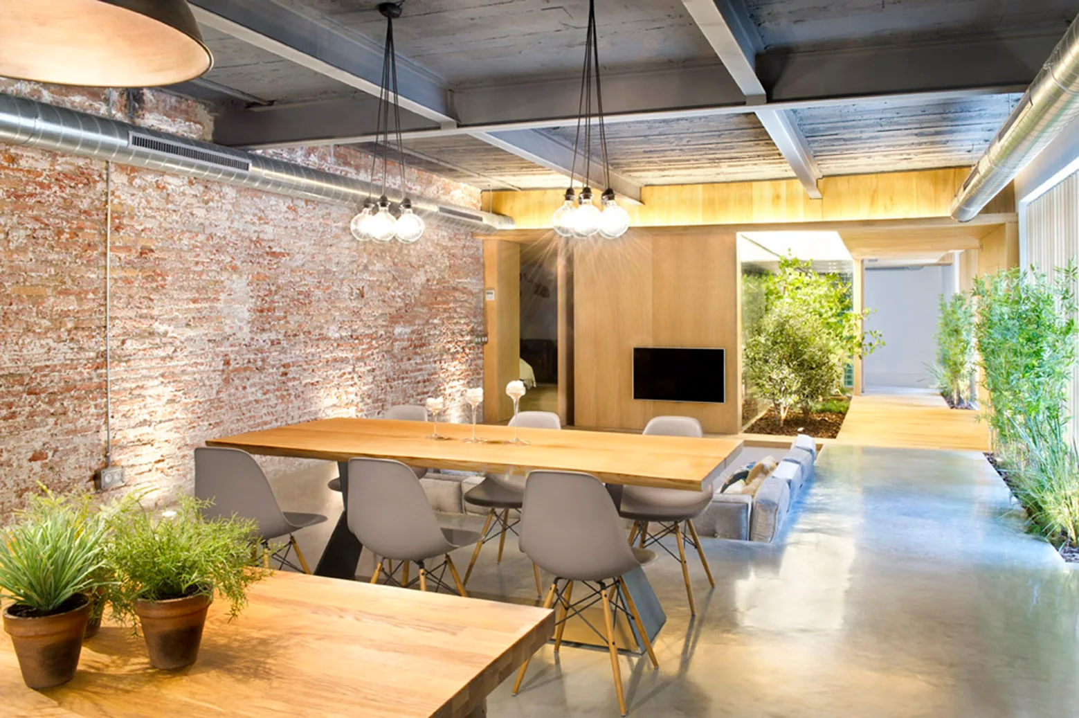 A long wooden dining table and grey chairs sit on a polished concrete floor beside a rustic exposed brick wall and an indoor garden area.