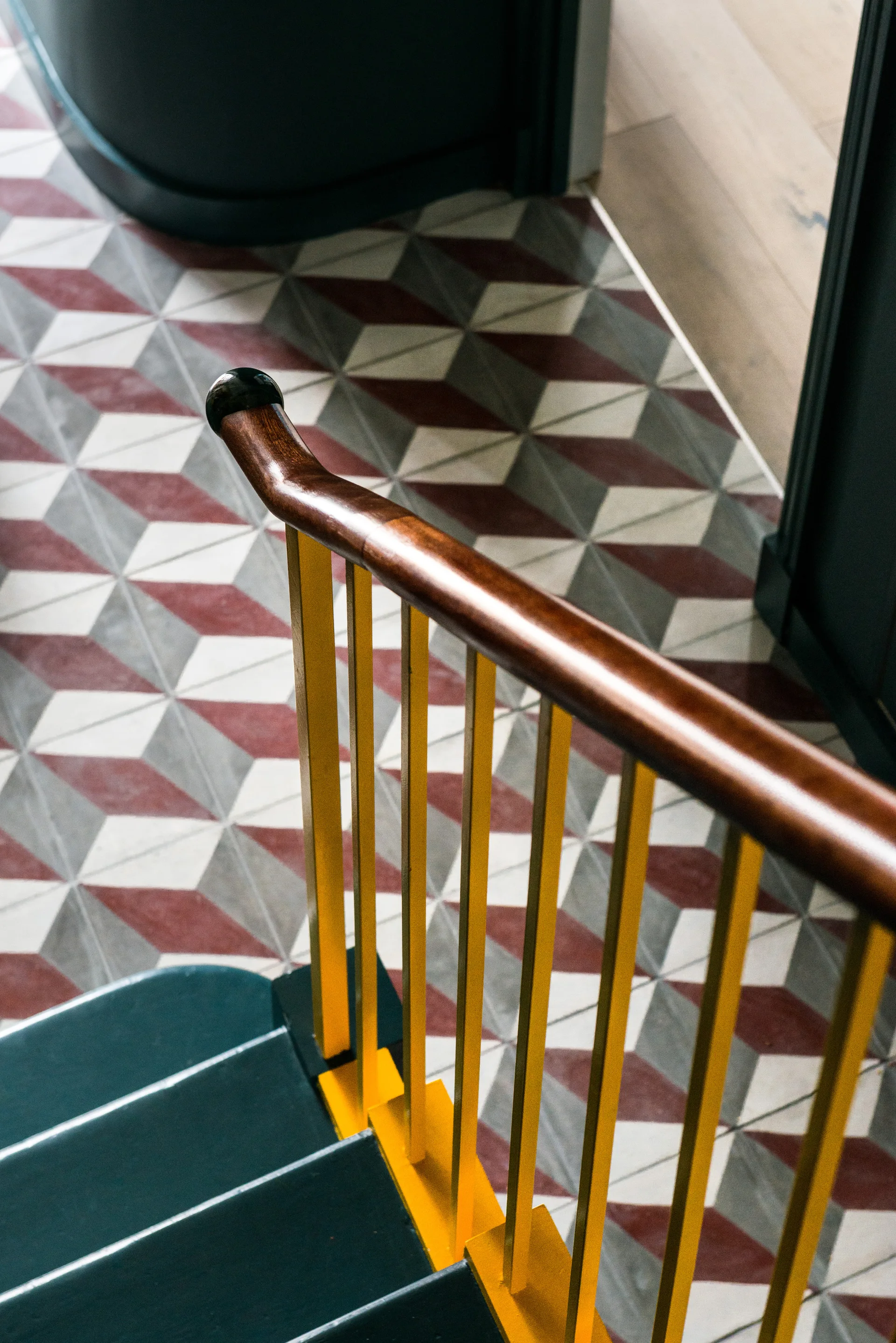 A staircase featuring dark teal steps, a polished wood handrail, and bright yellow balusters sits above a red, white, and grey geometric tiled floor.