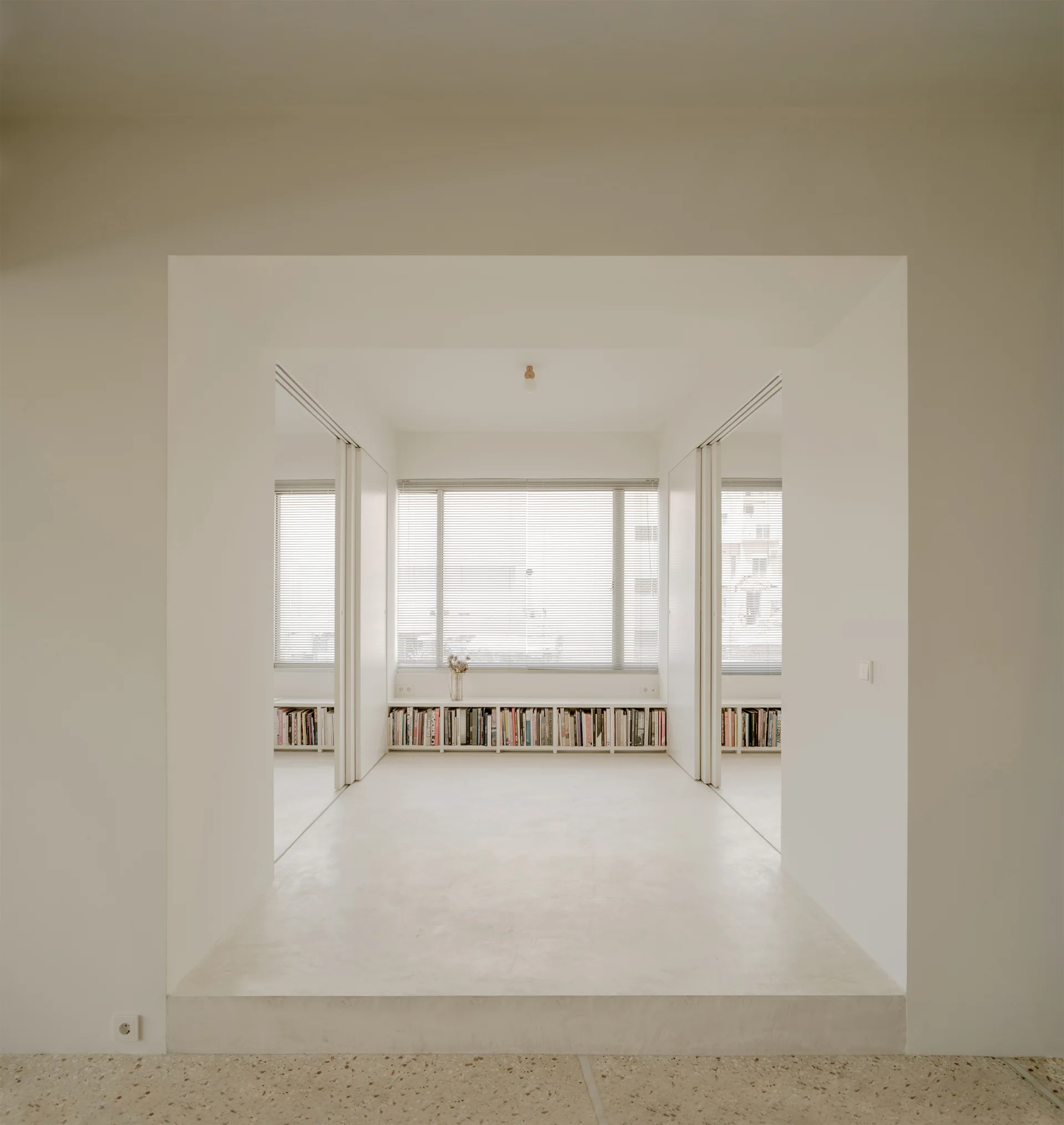 A minimalist off-white room features a raised floor accessed through a wide archway, with low bookshelves set beneath a large window with blinds.