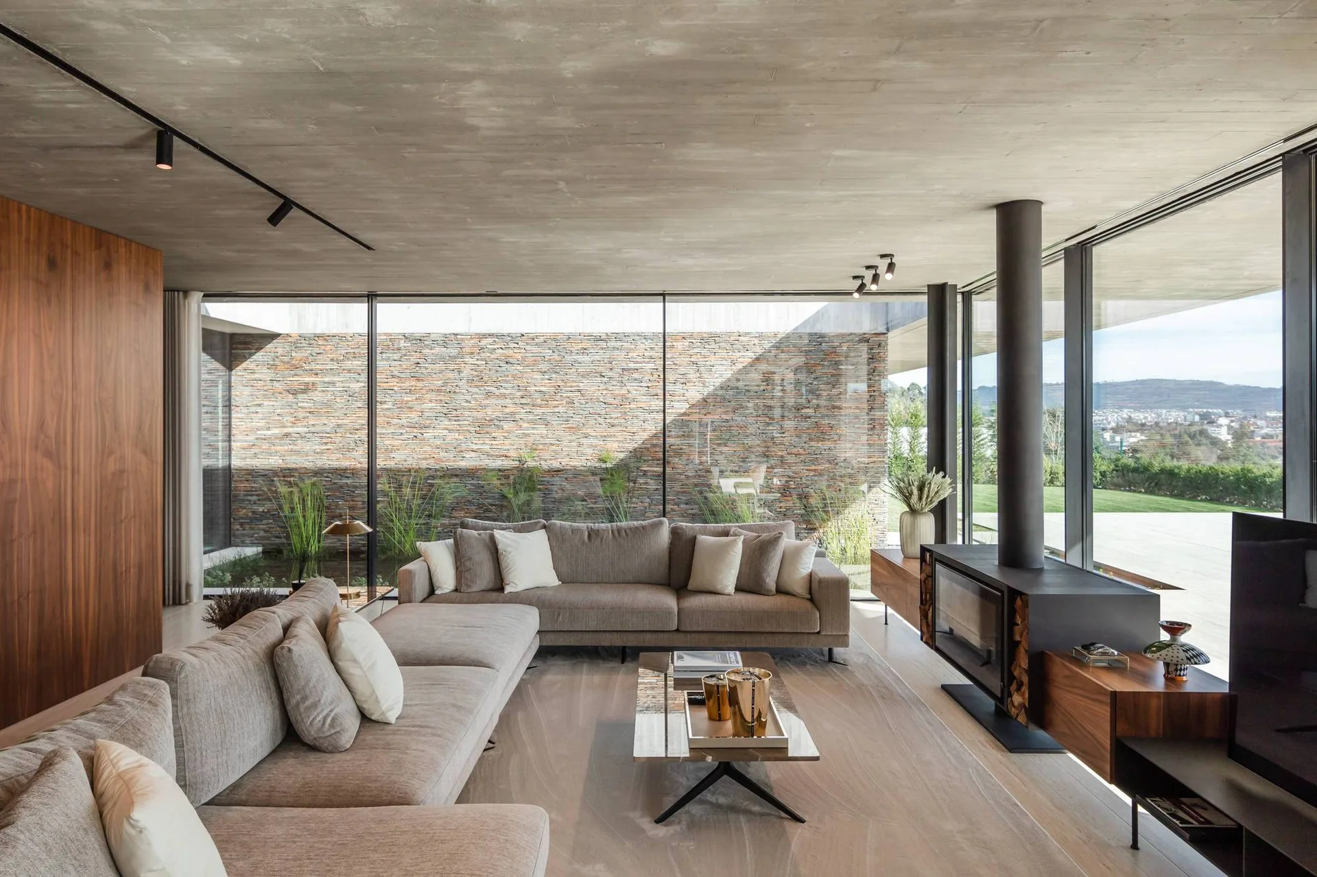 A modern living room features a concrete ceiling, large beige sectional sofas, a black freestanding fireplace, and floor-to-ceiling glass walls.