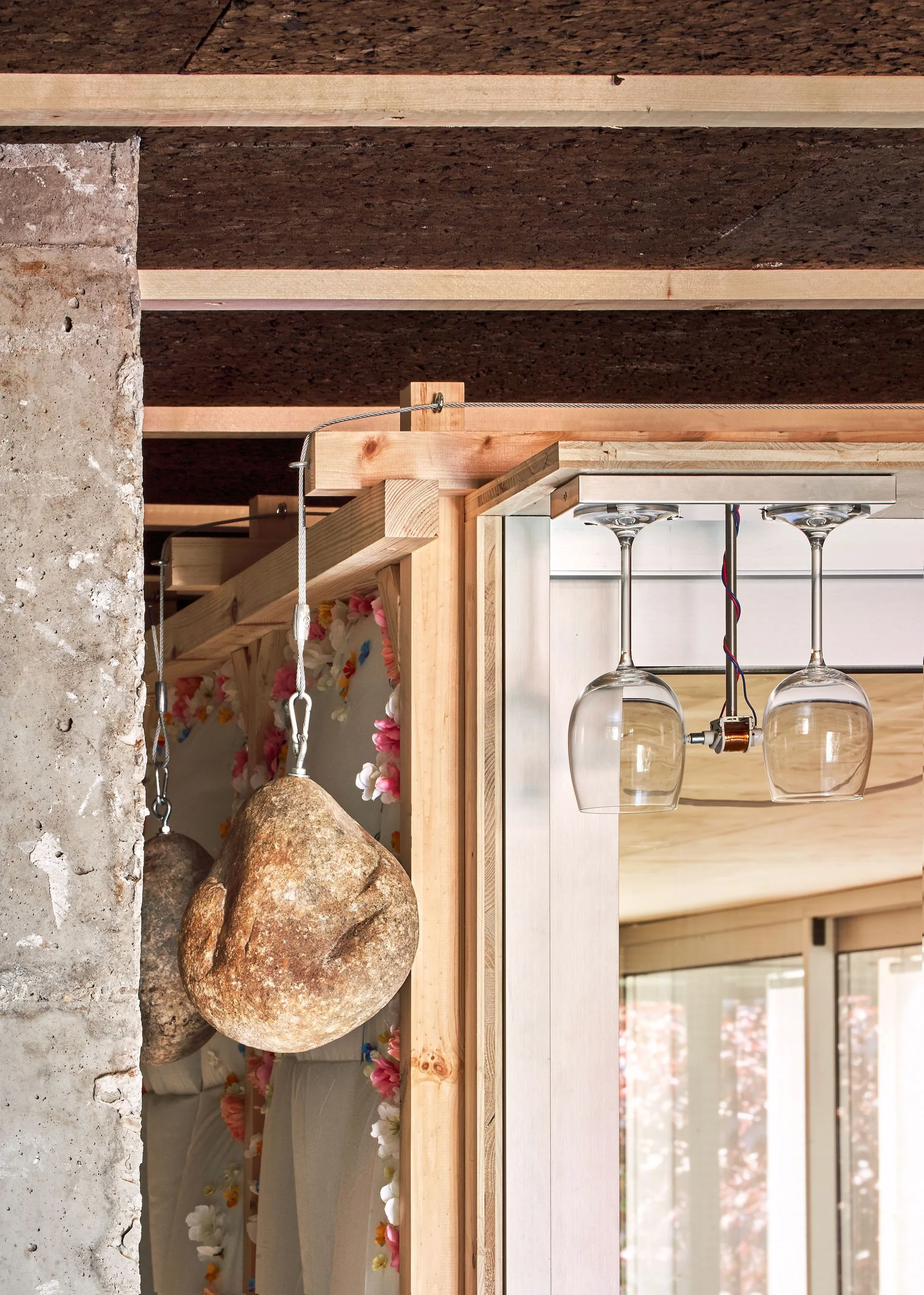 A close-up of an unfinished timber structure with hanging stone weights, suspended wine glasses, a textured cork ceiling, and a raw concrete column.