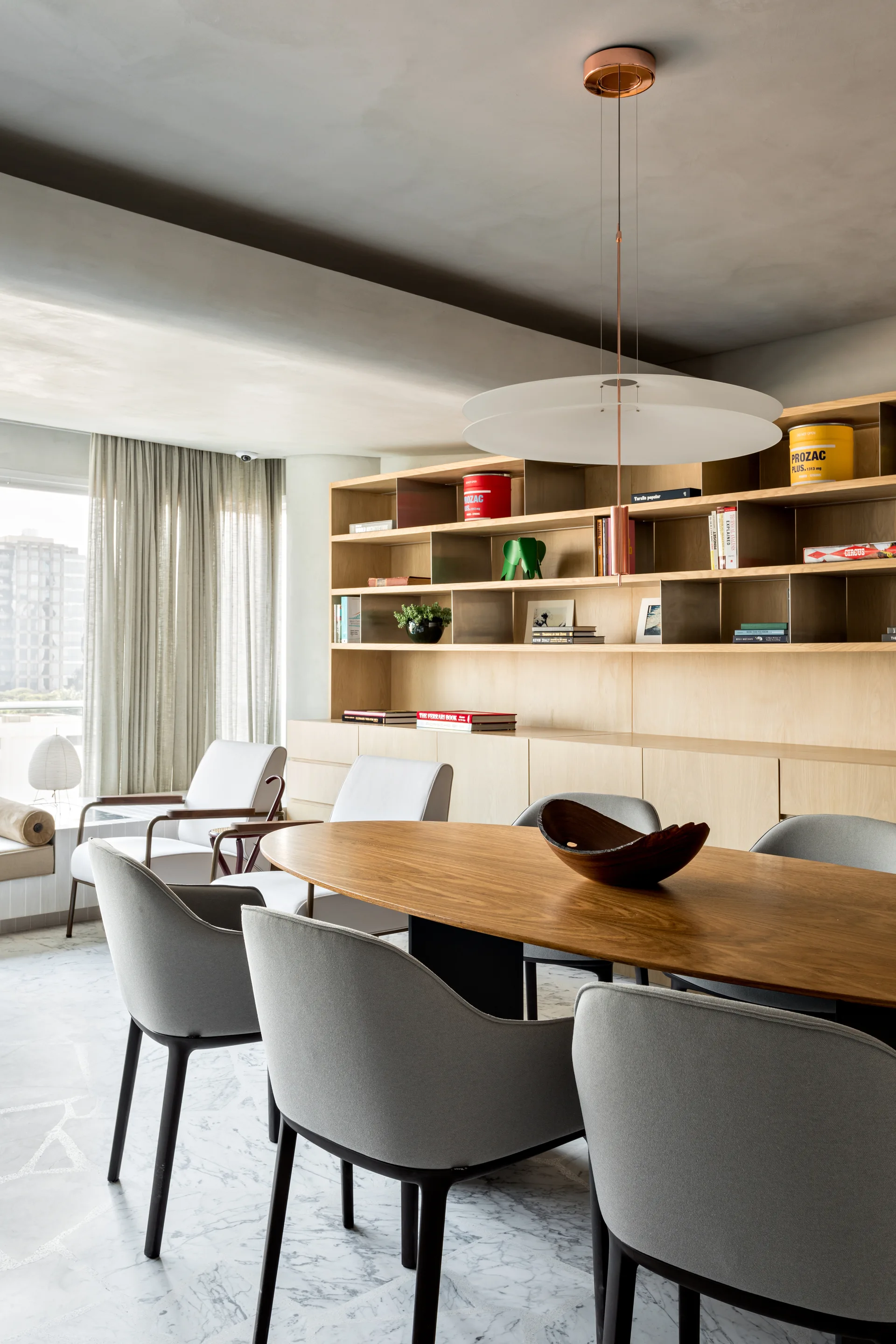 An oval wood dining table with grey chairs sits on a veined marble floor next to a large custom wooden shelving unit and a window with sheer curtains.