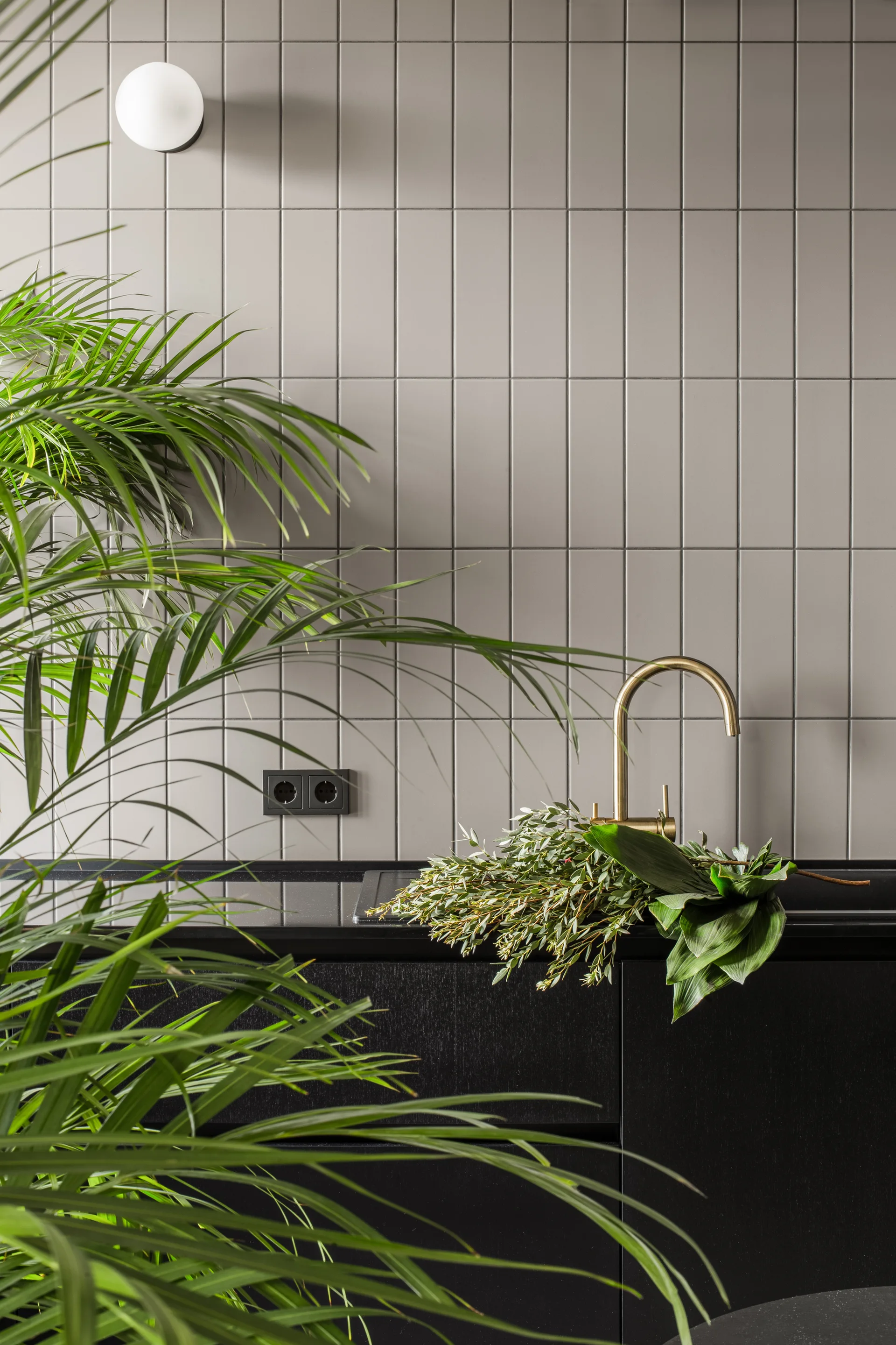 A close-up of a kitchen wall featuring a light grey vertical tile backsplash, a curved brass faucet on a black countertop, and fresh green foliage.