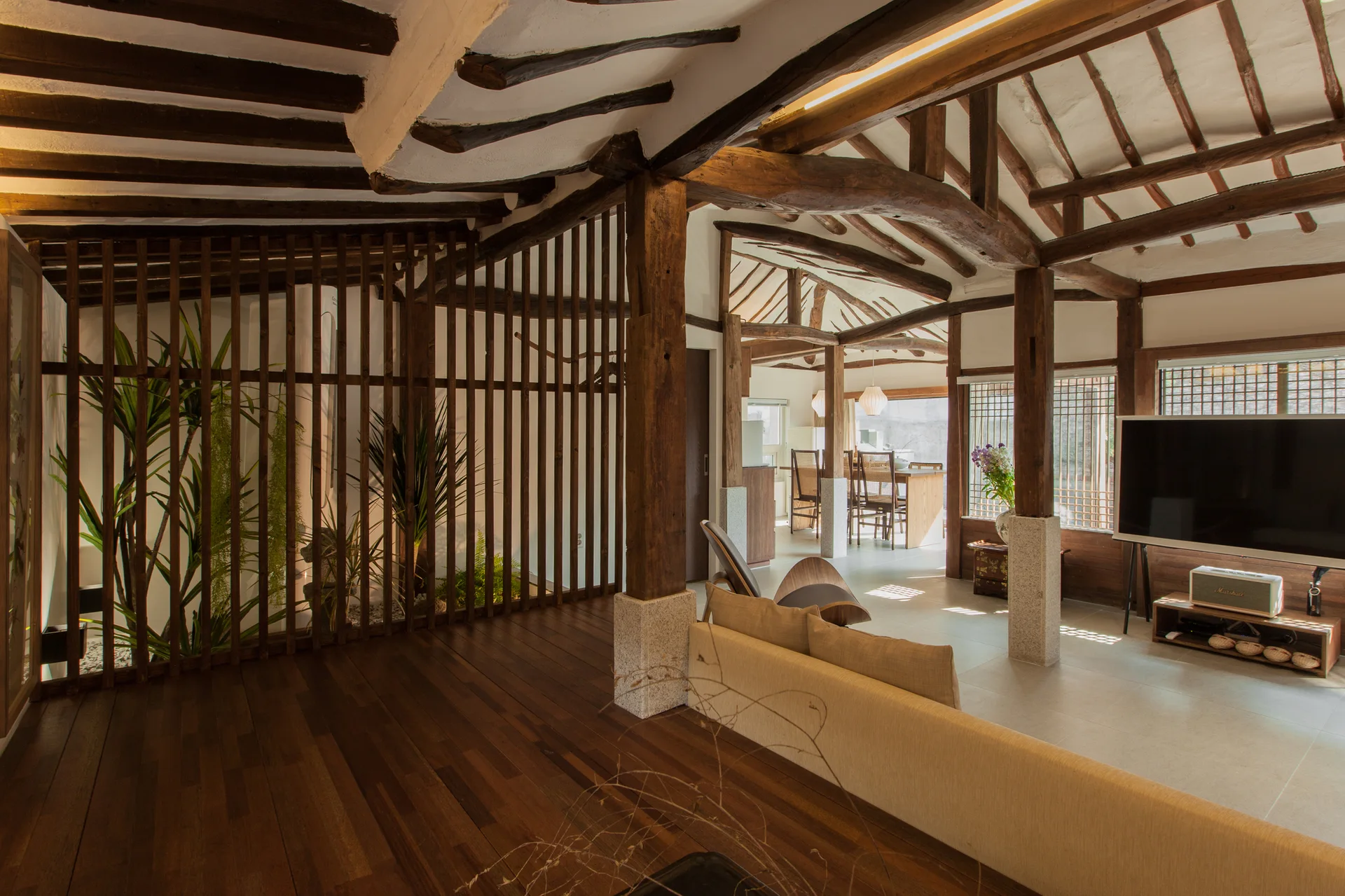 A minimalist living room features exposed dark wood ceiling beams, stone-base pillars, a beige sofa, and a vertical slatted wood screen.