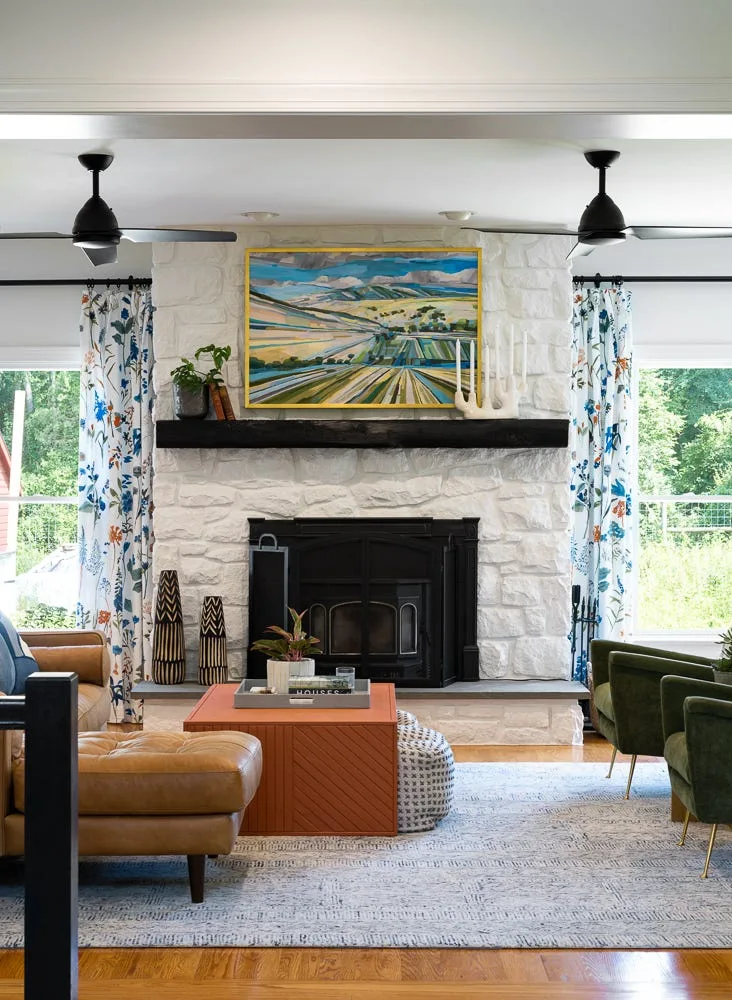 A transitional living room featuring a white stone fireplace with a rustic wood mantel, flanked by windows with blue and green floral curtains.