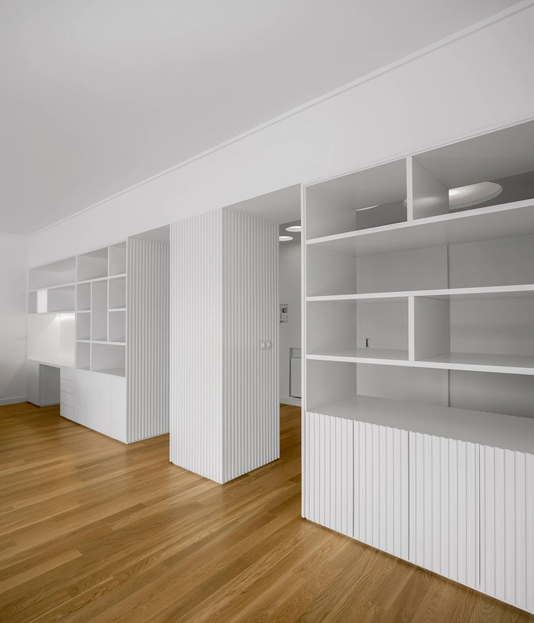 Custom white floor to ceiling shelving units with vertical slatted lower cabinets sit on natural hardwood flooring.