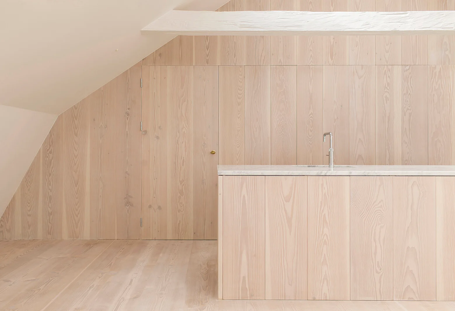 A minimalist attic kitchen features a light wood-clad island and matching wood paneled walls beneath a sloped ceiling with a white painted beam.