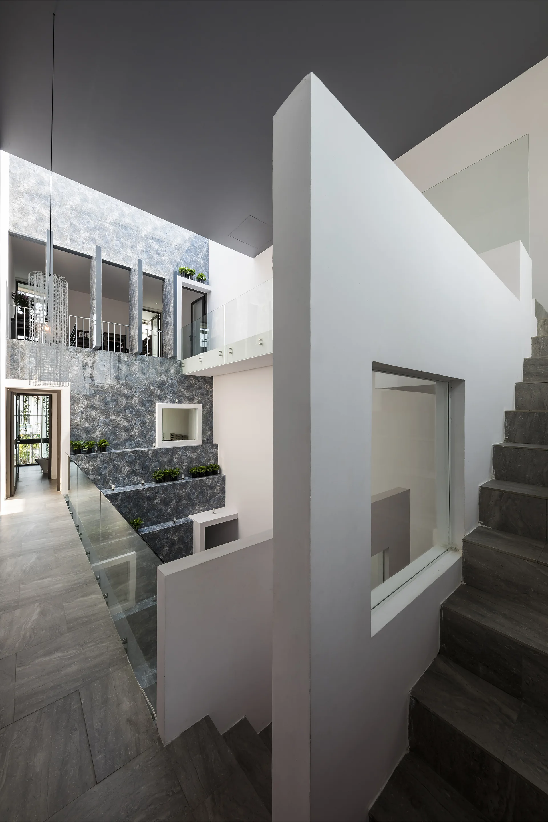 A multi-level modern interior stairwell features grey stone steps, a white partition wall with a square cutout window, and glass balustrades.