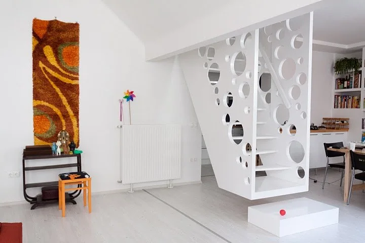 A modern white floating staircase with round cutouts stands in a bright room with light wood floors, a colorful tapestry, and a dining table.