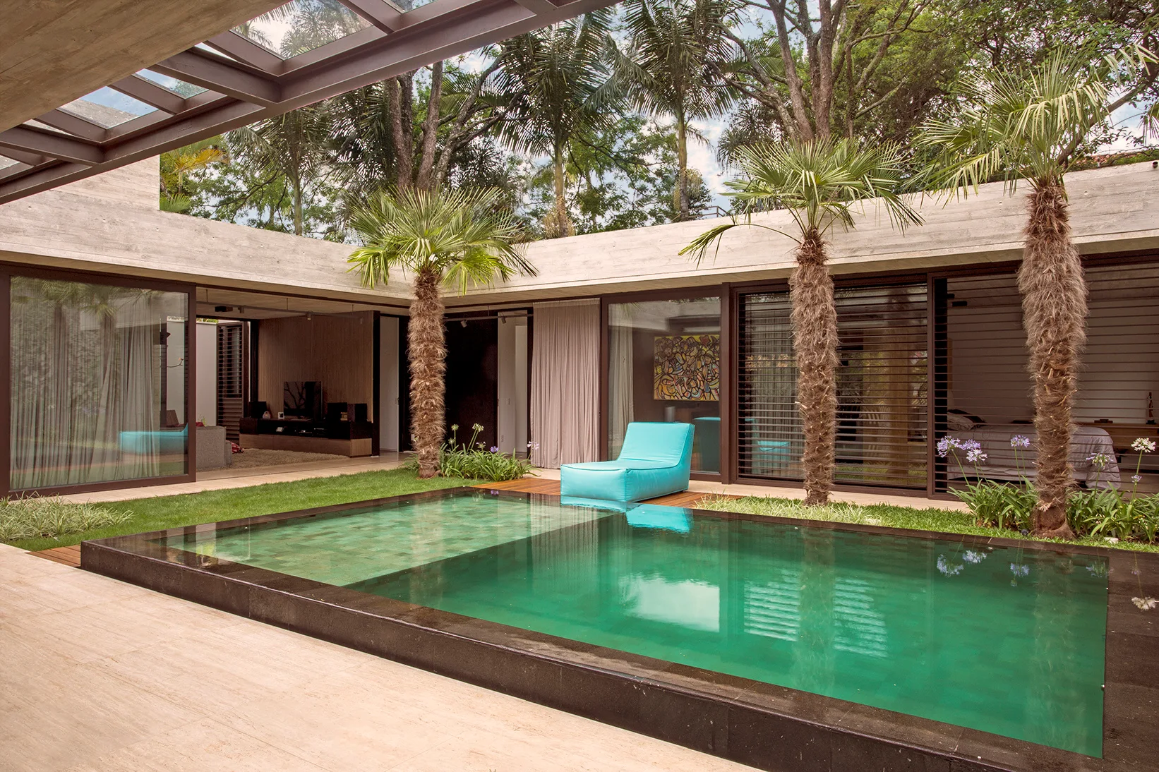 A modern open-air courtyard features a rectangular swimming pool framed by concrete walls, glass sliding doors, and two palm trees.