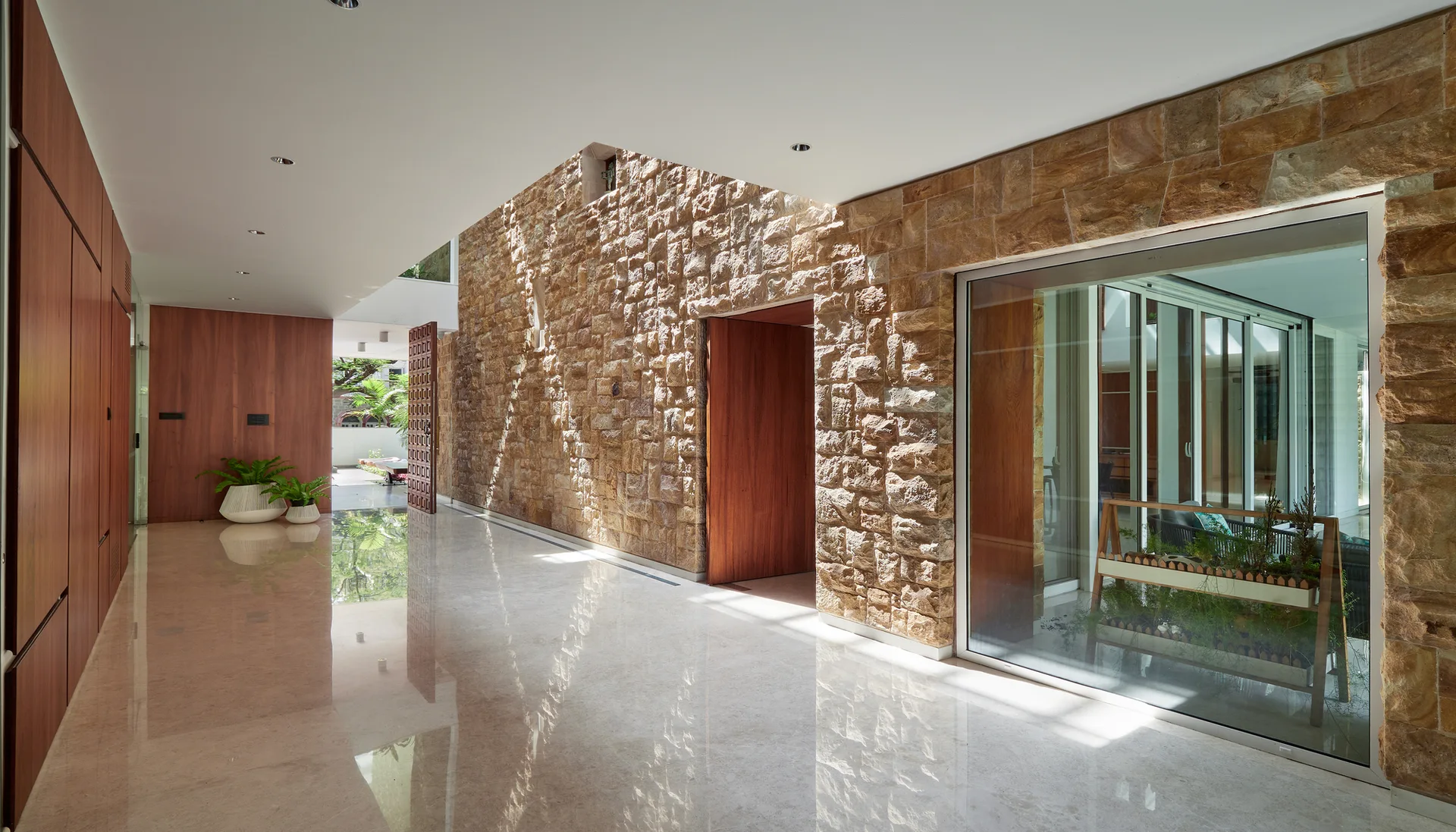 A wide hallway featuring a textured stone masonry wall, polished reflective floor, and warm wood wall panels with open doorways.