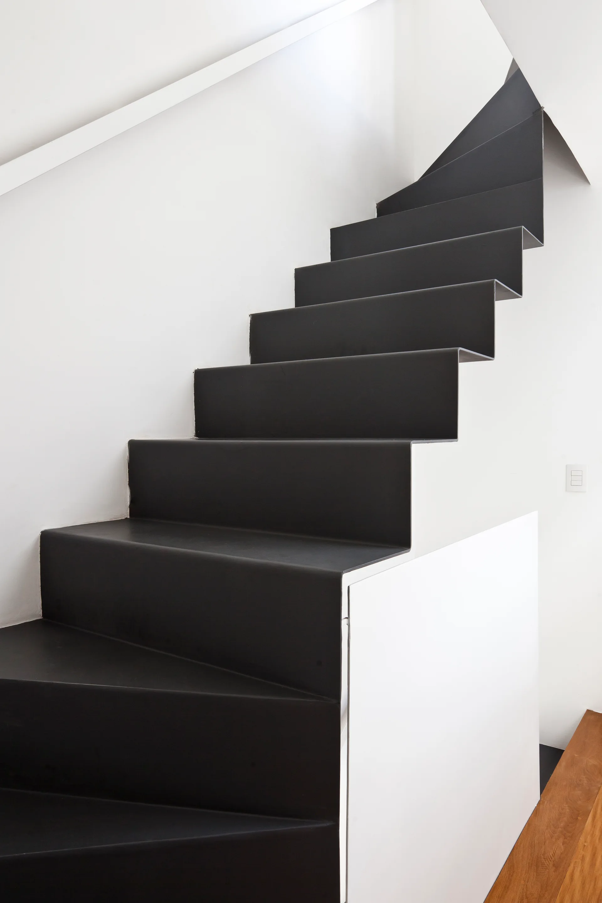 A minimalist black ribbon staircase ascends against white walls with integrated white storage at the base.