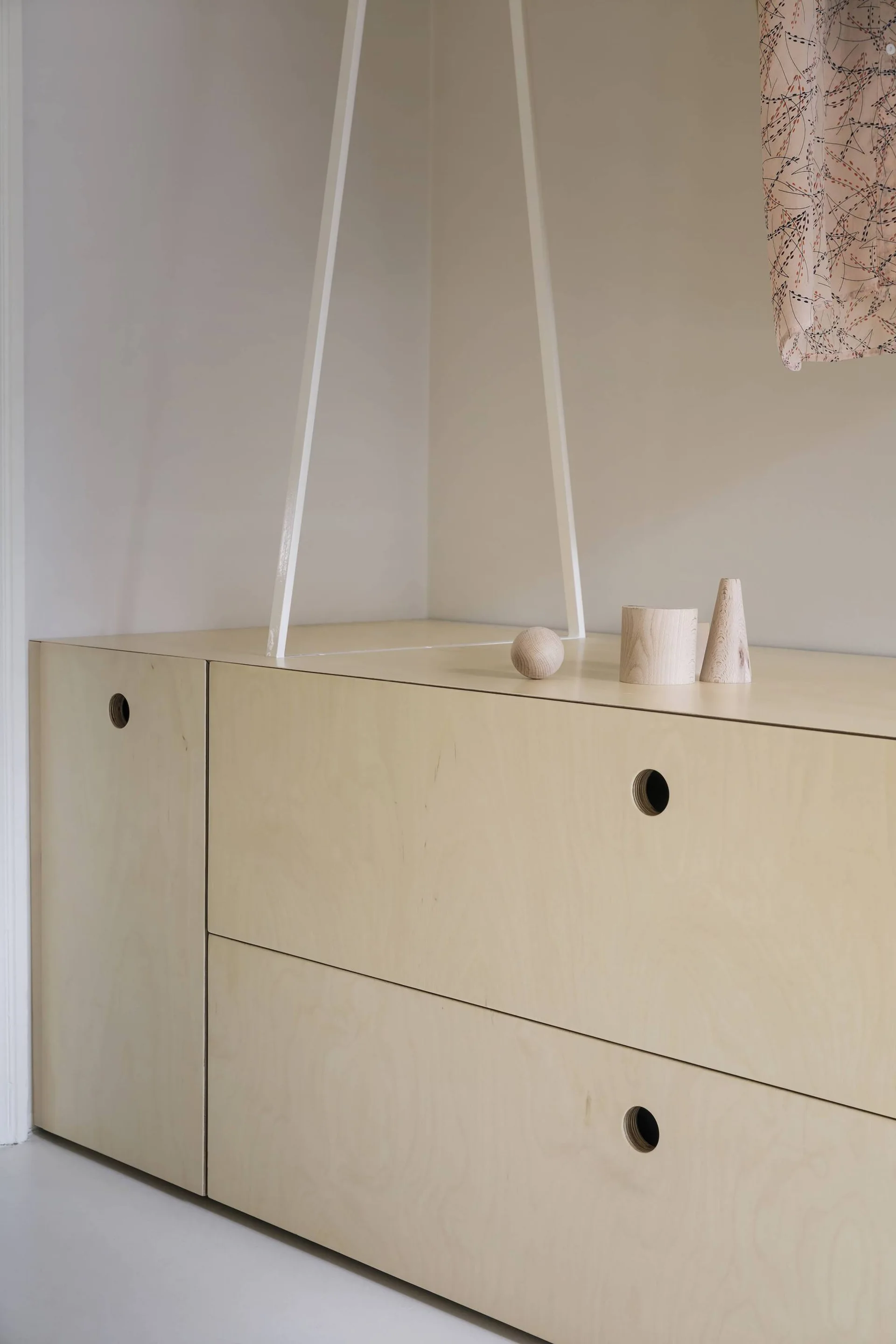 A light plywood storage cabinet with circular cut-out pulls stands against an off-white wall, topped with small geometric wooden shapes.
