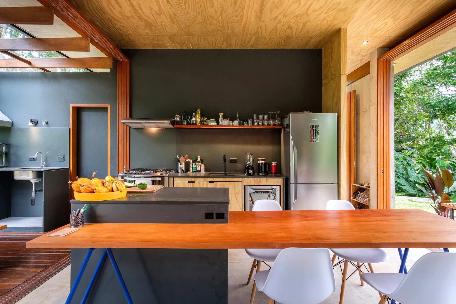 A modern open kitchen features a plywood ceiling, a dark gray island connected to a wooden dining table, and white chairs.