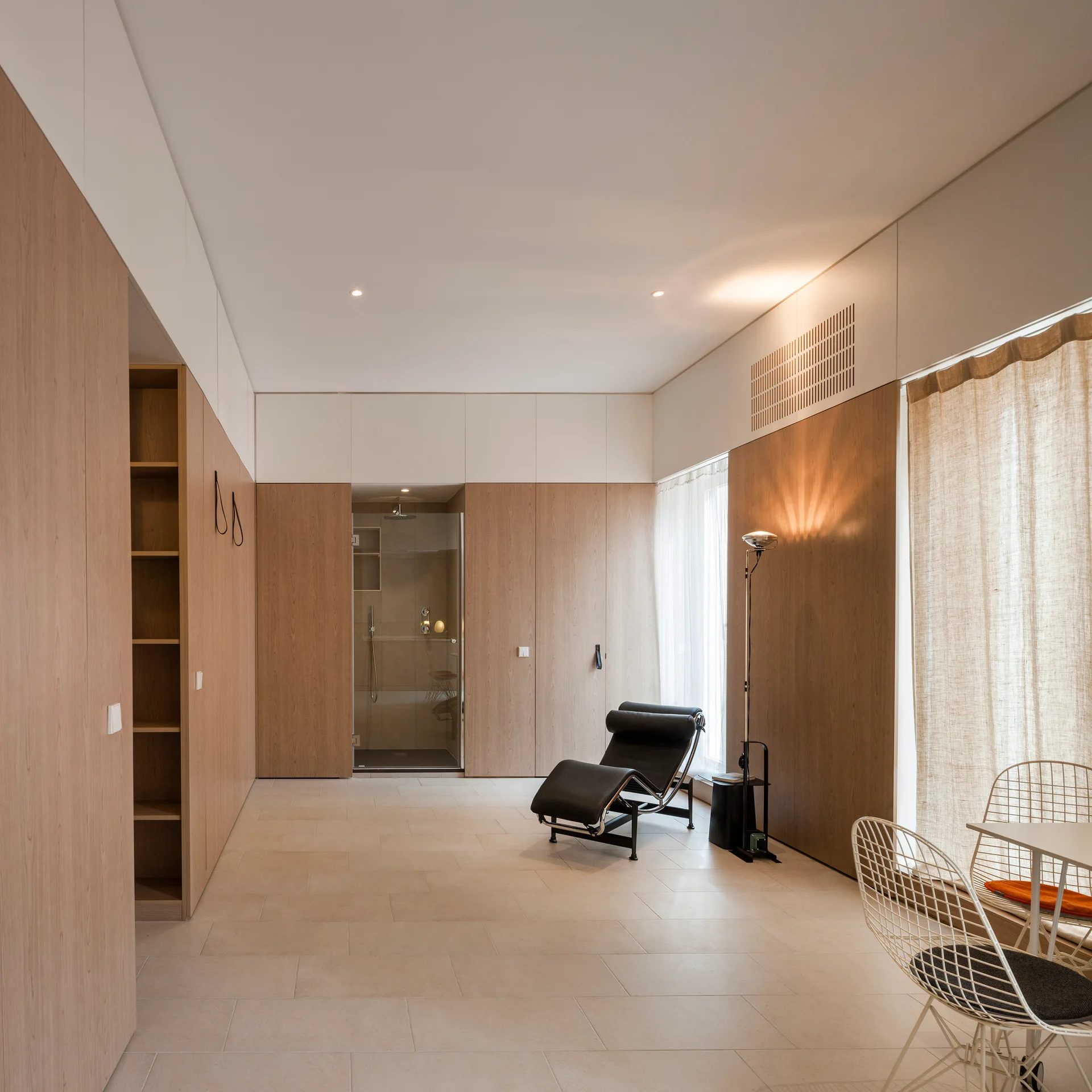 A minimalist bedroom features seamless light wood wall cabinetry, a black chaise lounge chair, a glass shower stall, and large format floor tiles.