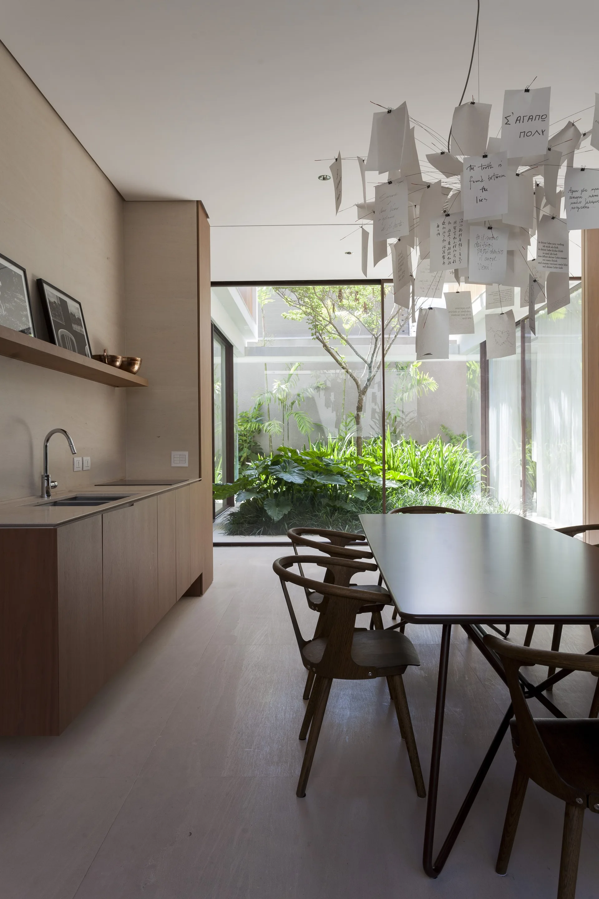 A minimalist dining room features a dark wood table, wood cabinetry, and a paper note pendant light next to large windows facing a green courtyard.