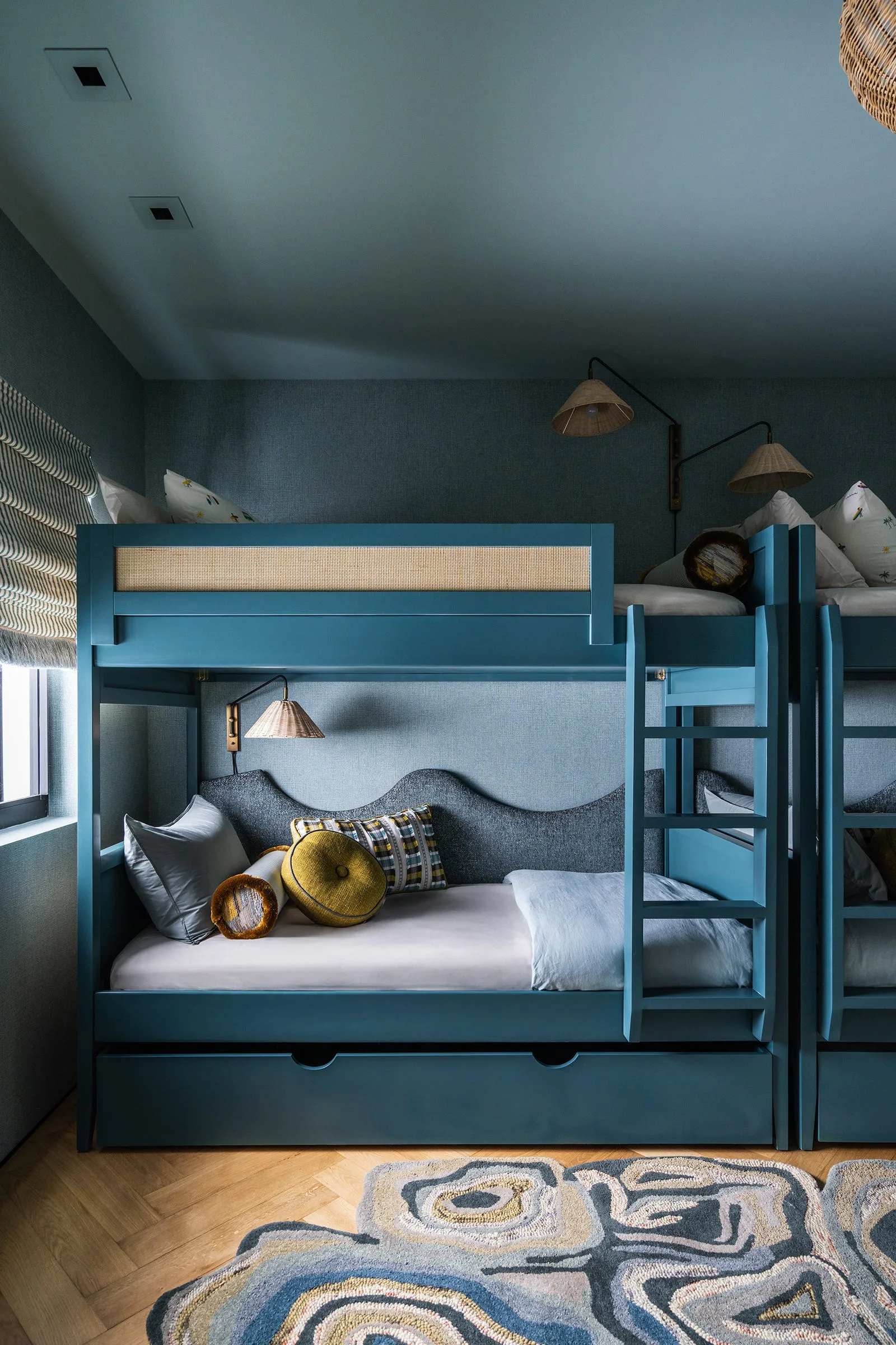 Blue painted wooden bunk beds feature woven rattan guard rails, a scalloped grey headboard, and wicker sconces against matching textured wallpaper.