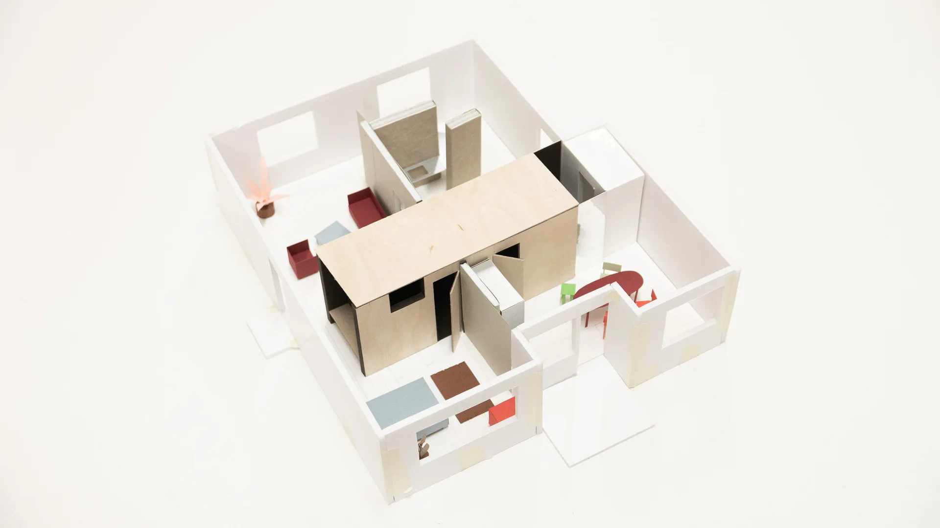 A physical scale model of an apartment floor plan featuring white walls, a central light wood structure, and miniature red and blue furniture.