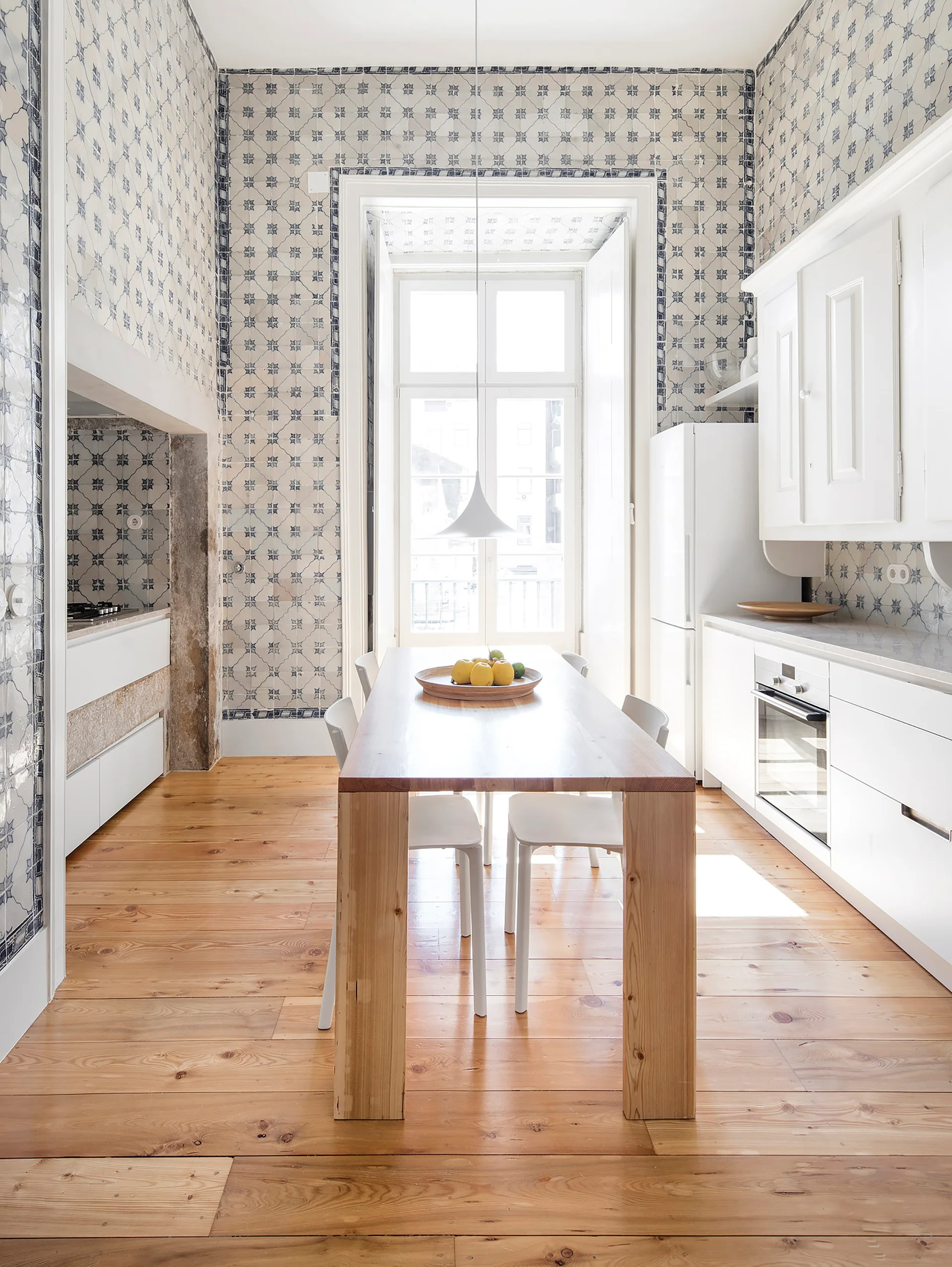 A bright kitchen features a wooden dining table and chairs, white modern cabinets, and walls covered in blue and white patterned tiles.