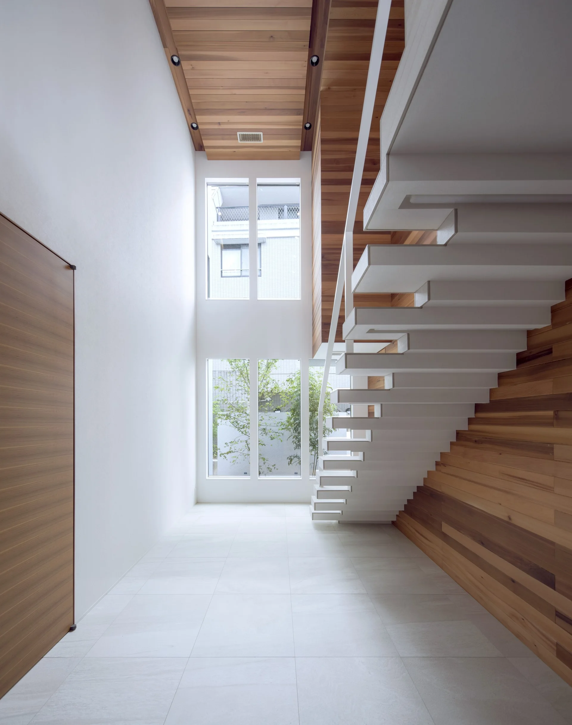 A double-height entryway featuring a white floating staircase against a warm wood-paneled wall, with large windows overlooking trees.