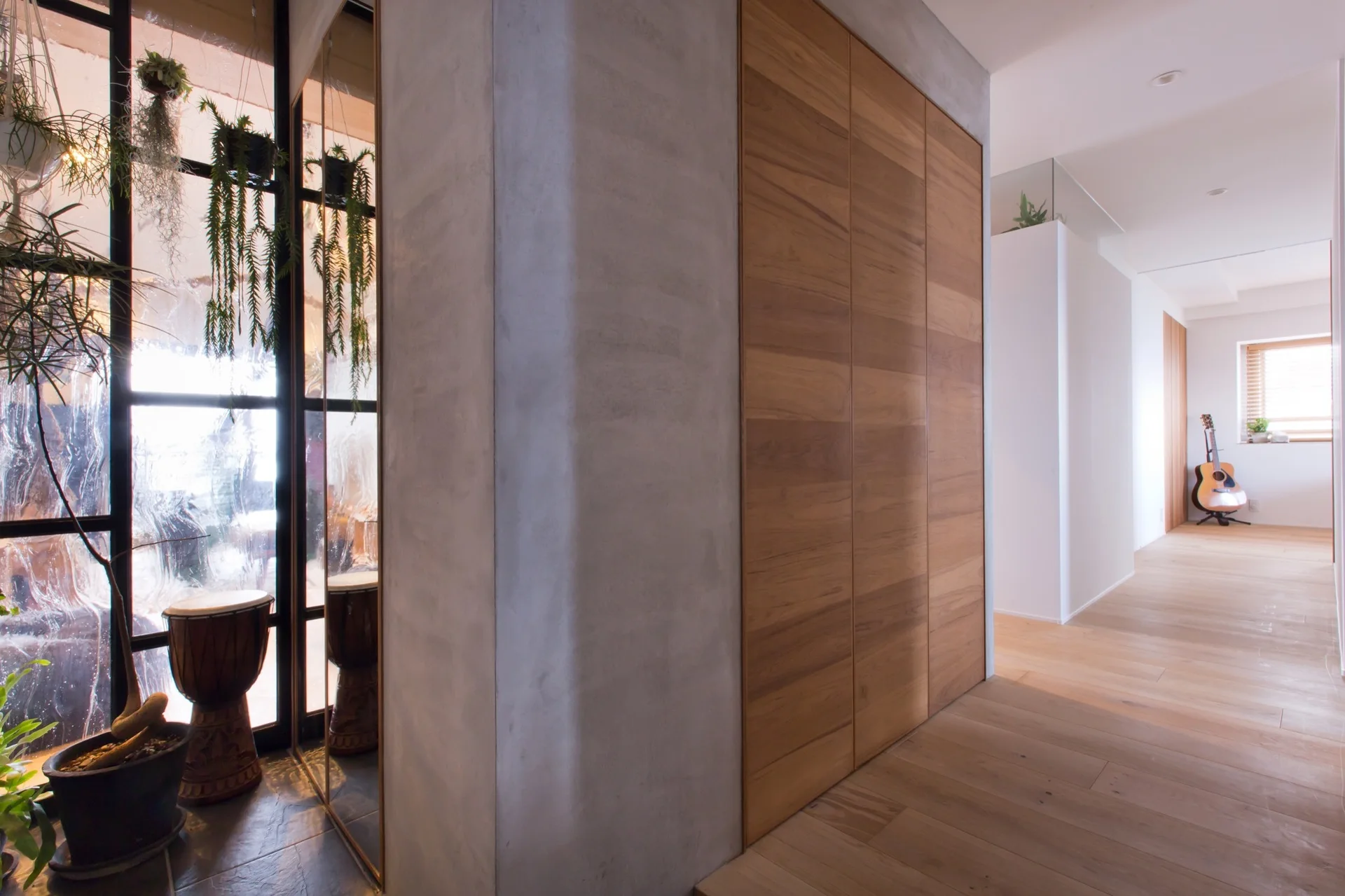A residential hallway features a concrete pillar next to wood-paneled closet doors, with a textured glass partition and hanging plants on the left.