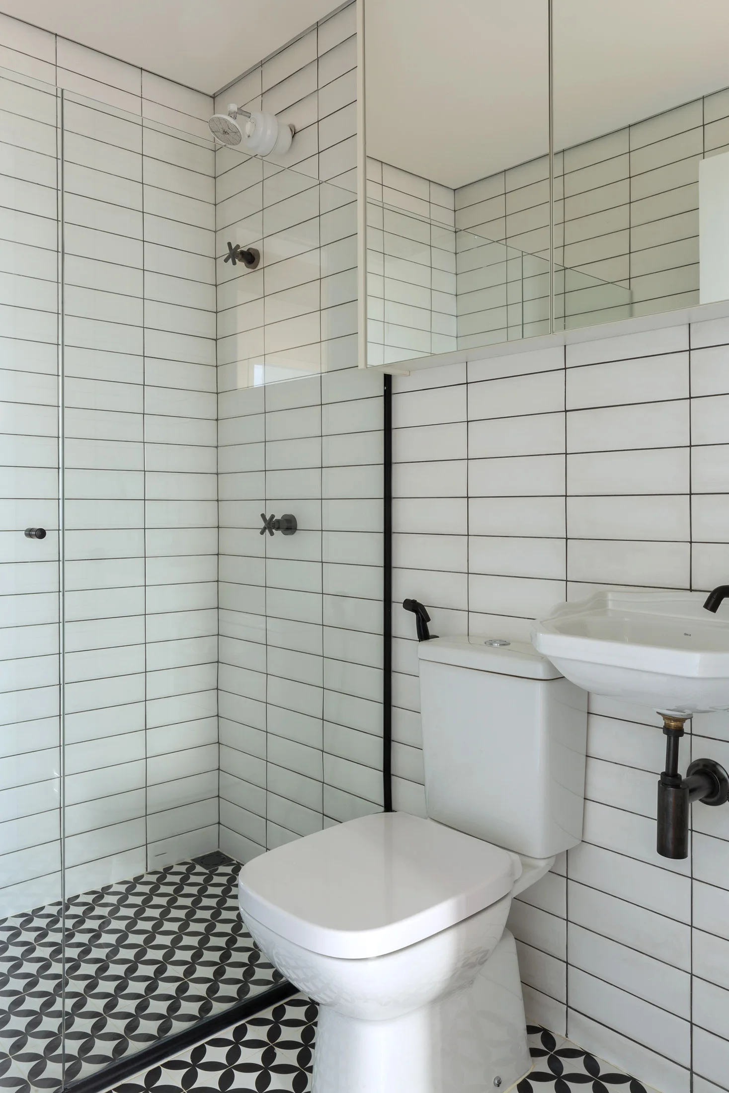 A modern bathroom features white stacked wall tiles, a black and white geometric patterned floor, a wall-mounted sink, and matte black hardware.