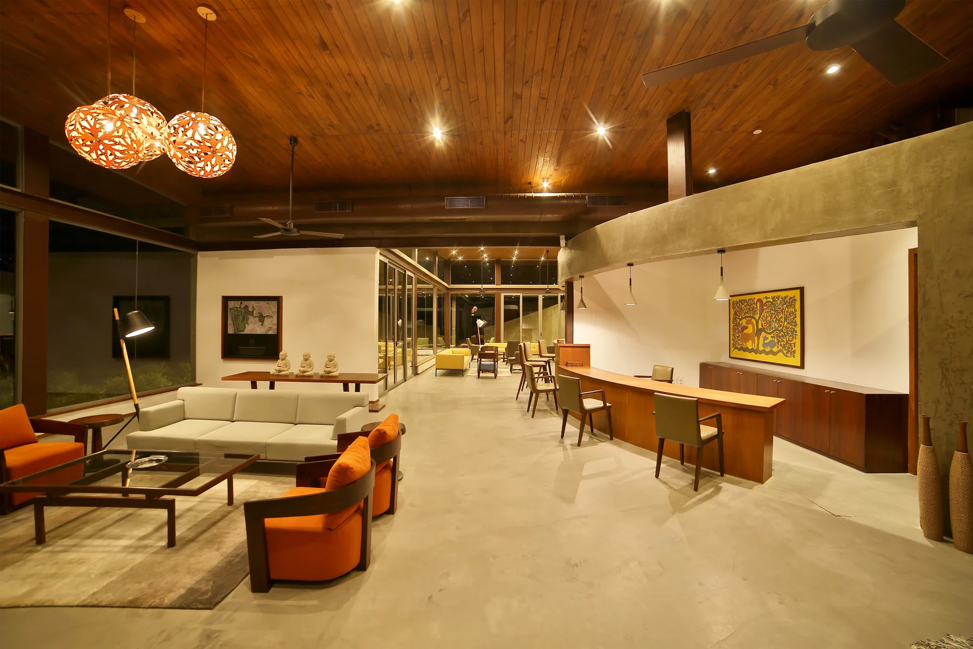 A spacious open-plan room with a wood plank ceiling, polished concrete floors, a light gray sofa, orange armchairs, and a long wooden bar counter.