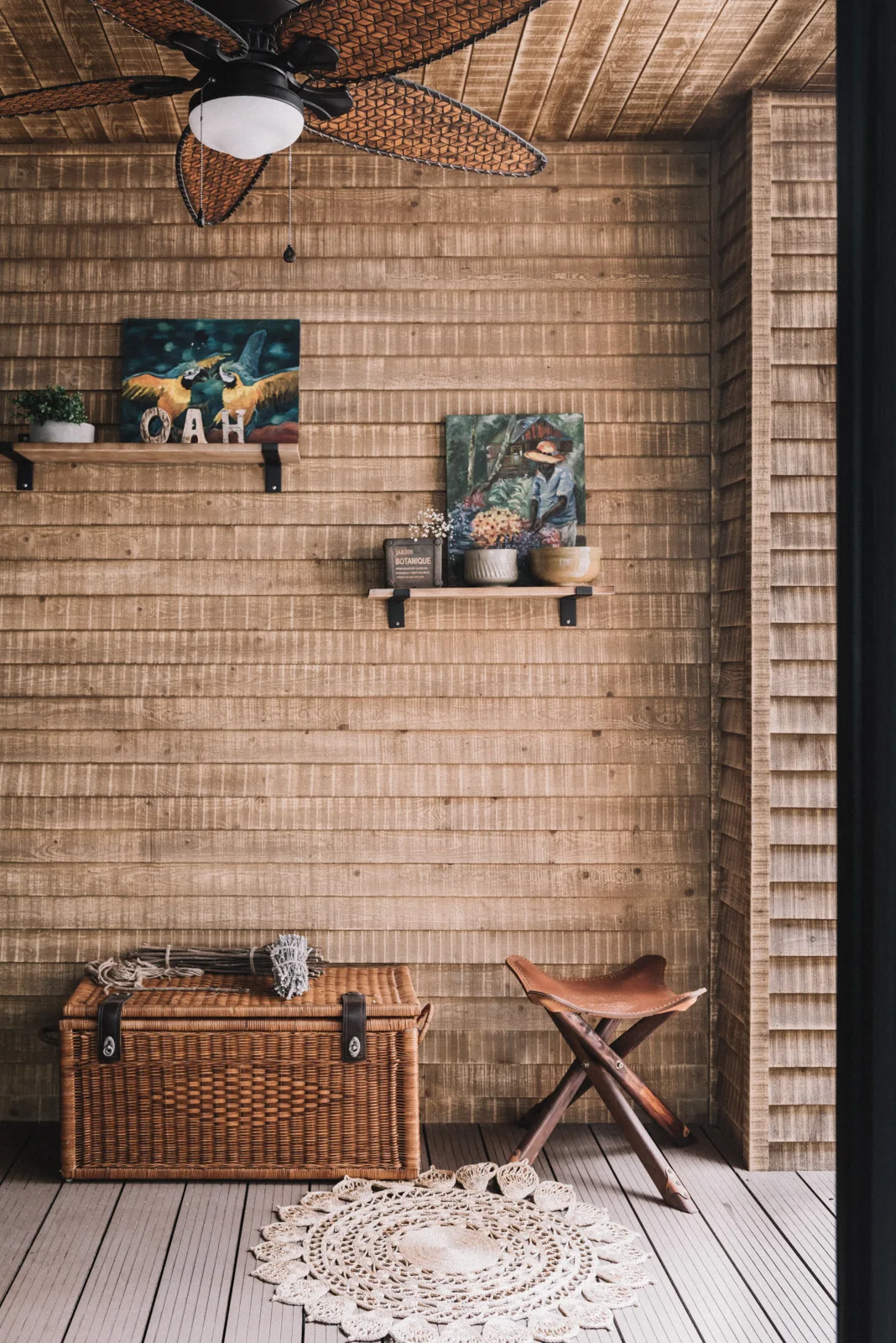 A covered wooden porch features natural horizontal wall paneling, a wicker storage trunk, a leather folding stool, and a decorative round woven rug.