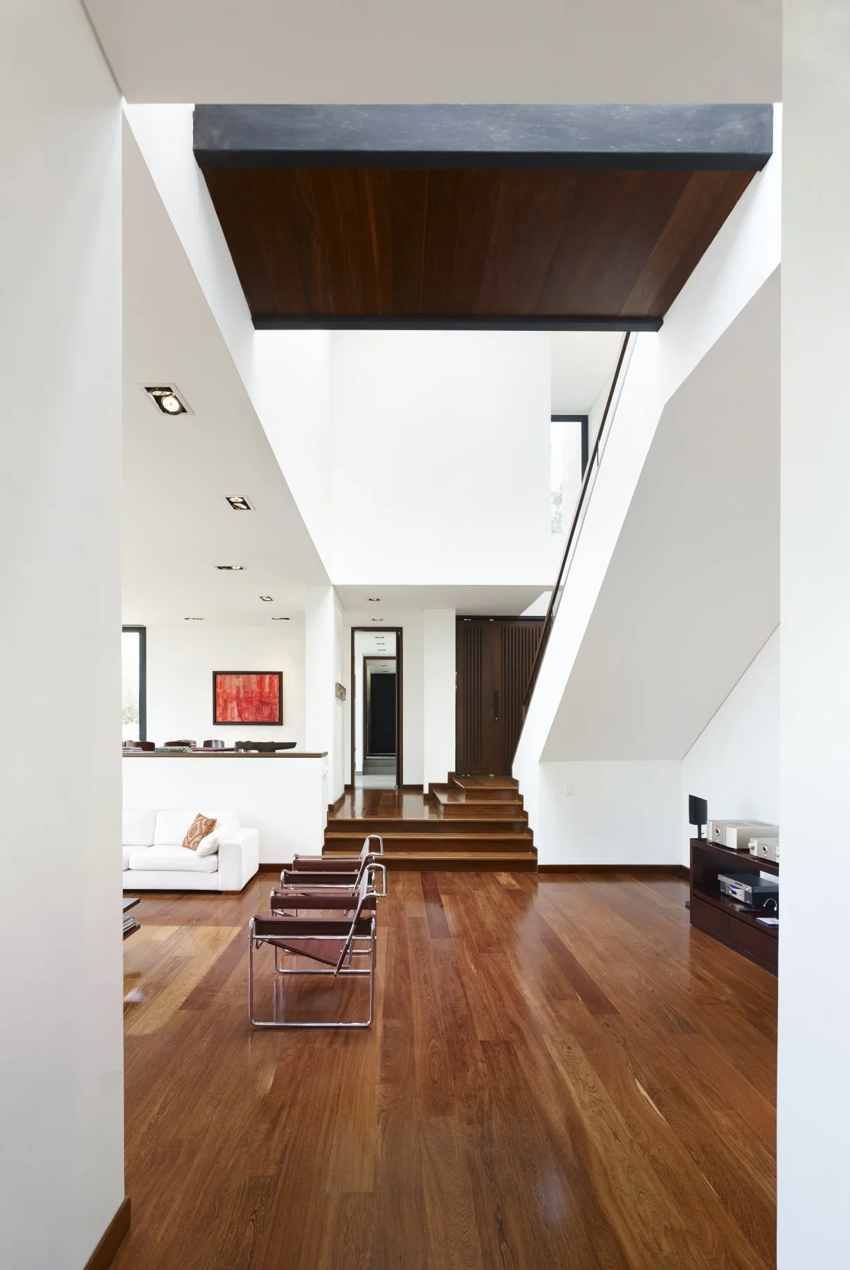 A spacious modern living area with white walls, warm hardwood floors, a skylight atrium, and two leather sling chairs.