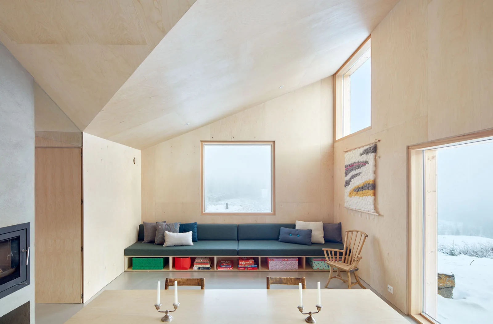 A minimalist cabin interior featuring plywood paneled walls, a sloped ceiling, and a green built-in storage bench next to a window overlooking snow.