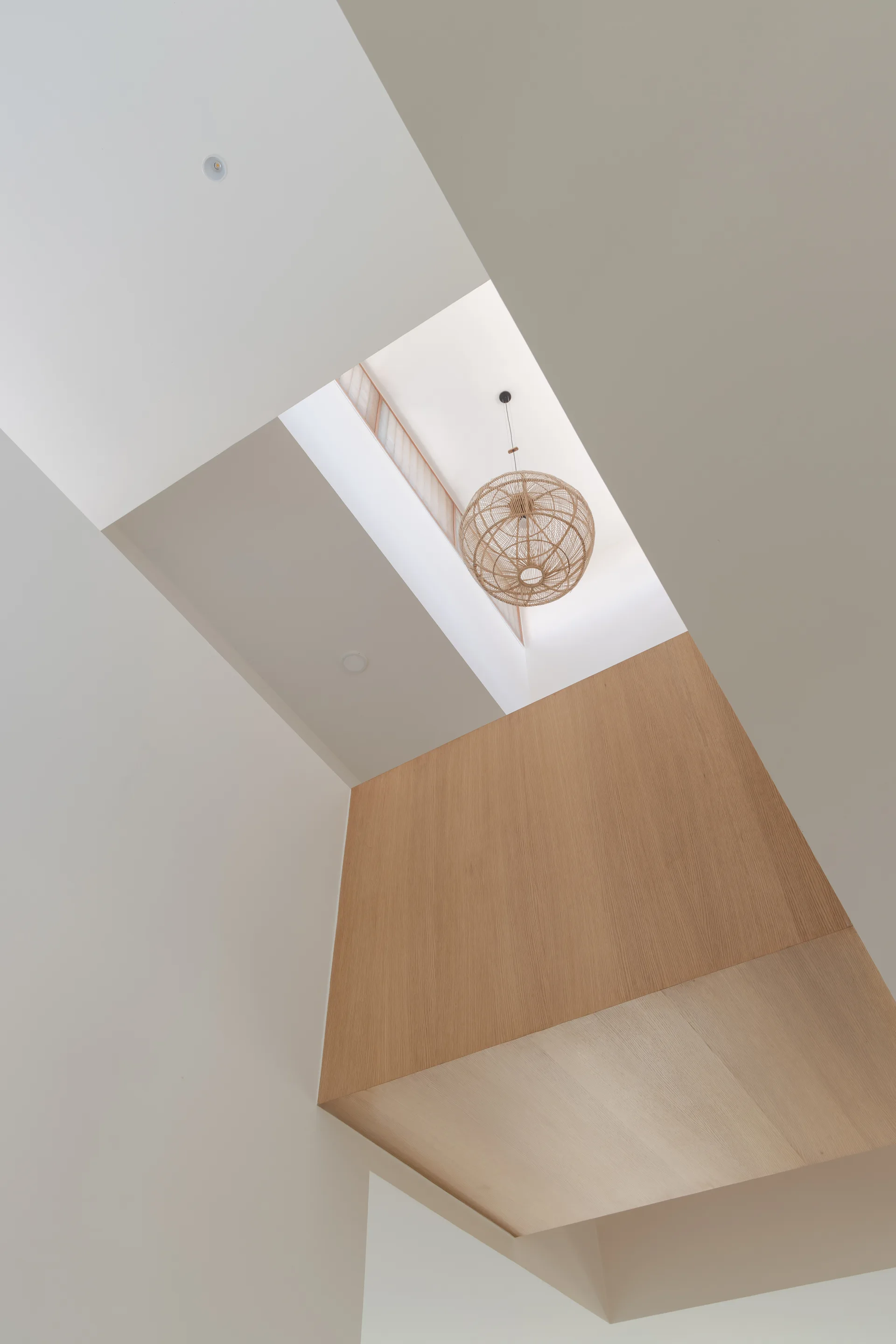 A low-angle view looking up into a high white ceiling void featuring a light wood accent block and a spherical woven pendant light.