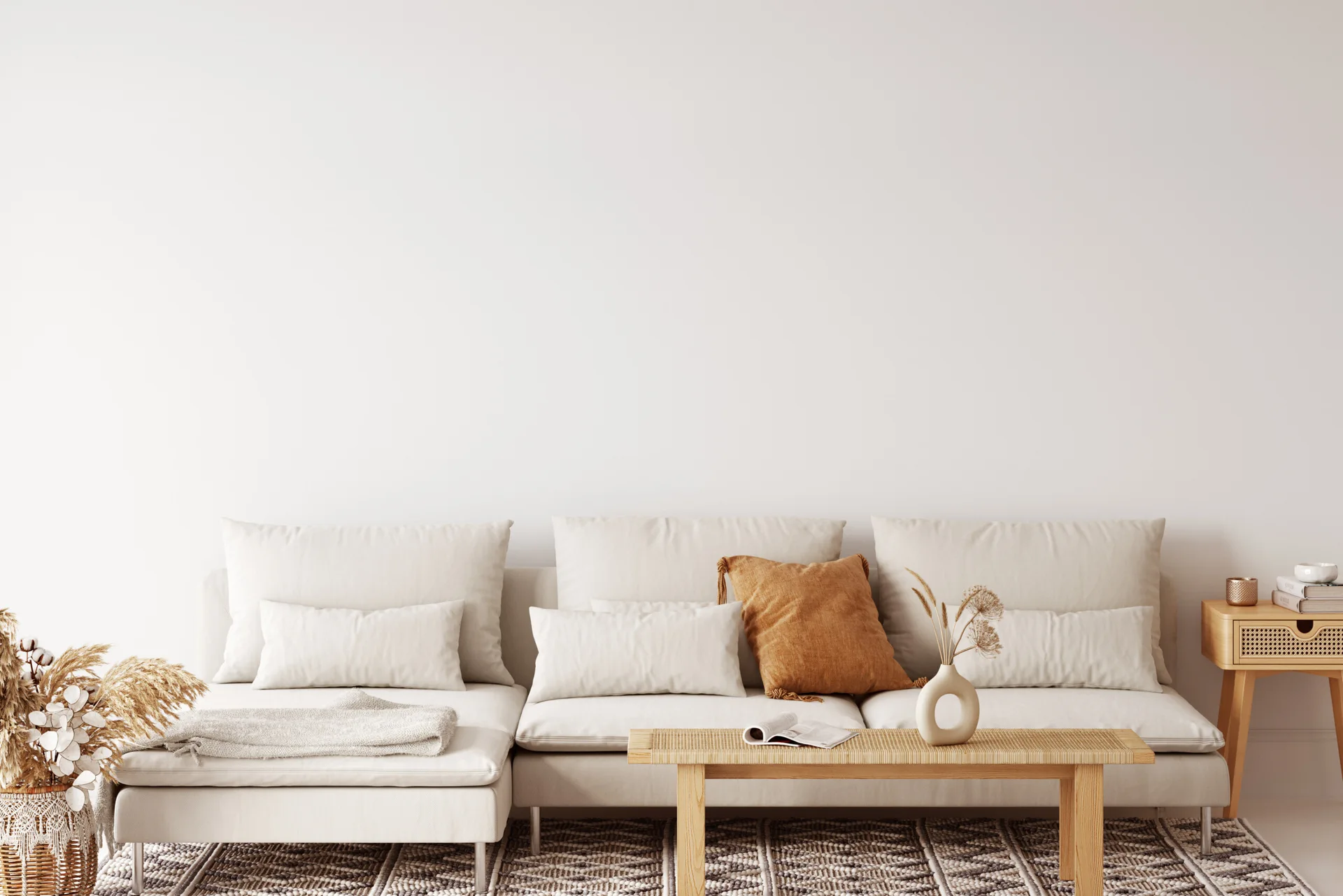 A minimalist cream sectional sofa paired with a low rattan coffee table and an ochre accent pillow against a white wall.