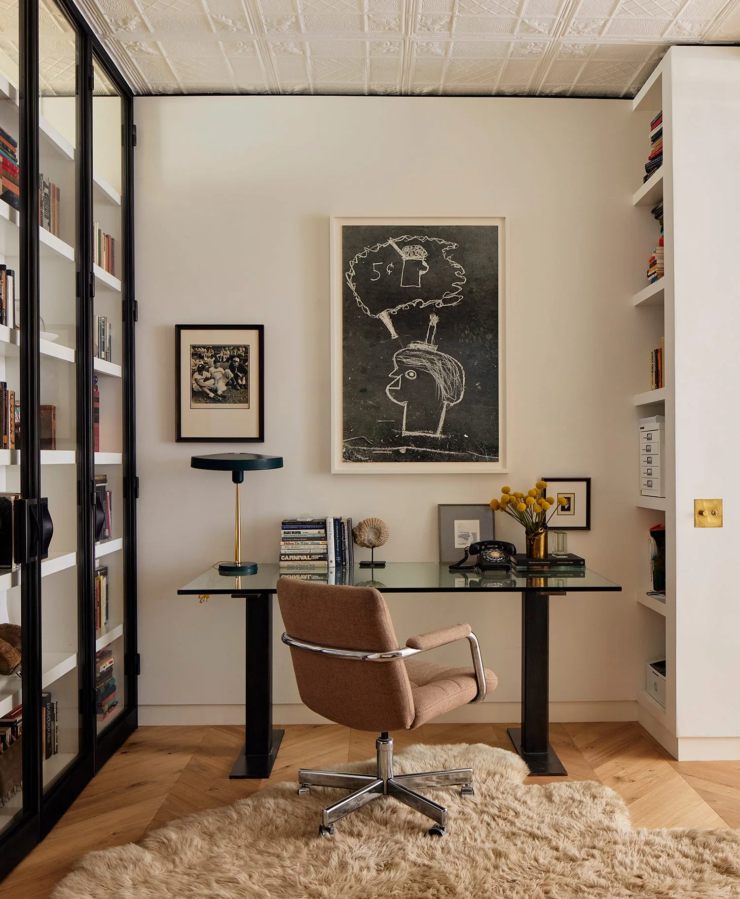 A home office featuring a glass-top desk, a tan swivel chair on a shag rug, herringbone wood floors, and built-in white bookshelves.