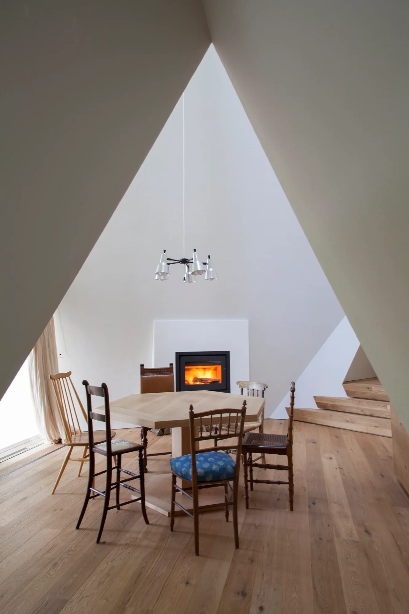 A hexagonal wooden dining table and mismatched chairs sit beneath a high, triangular white ceiling with a fireplace burning in the background.