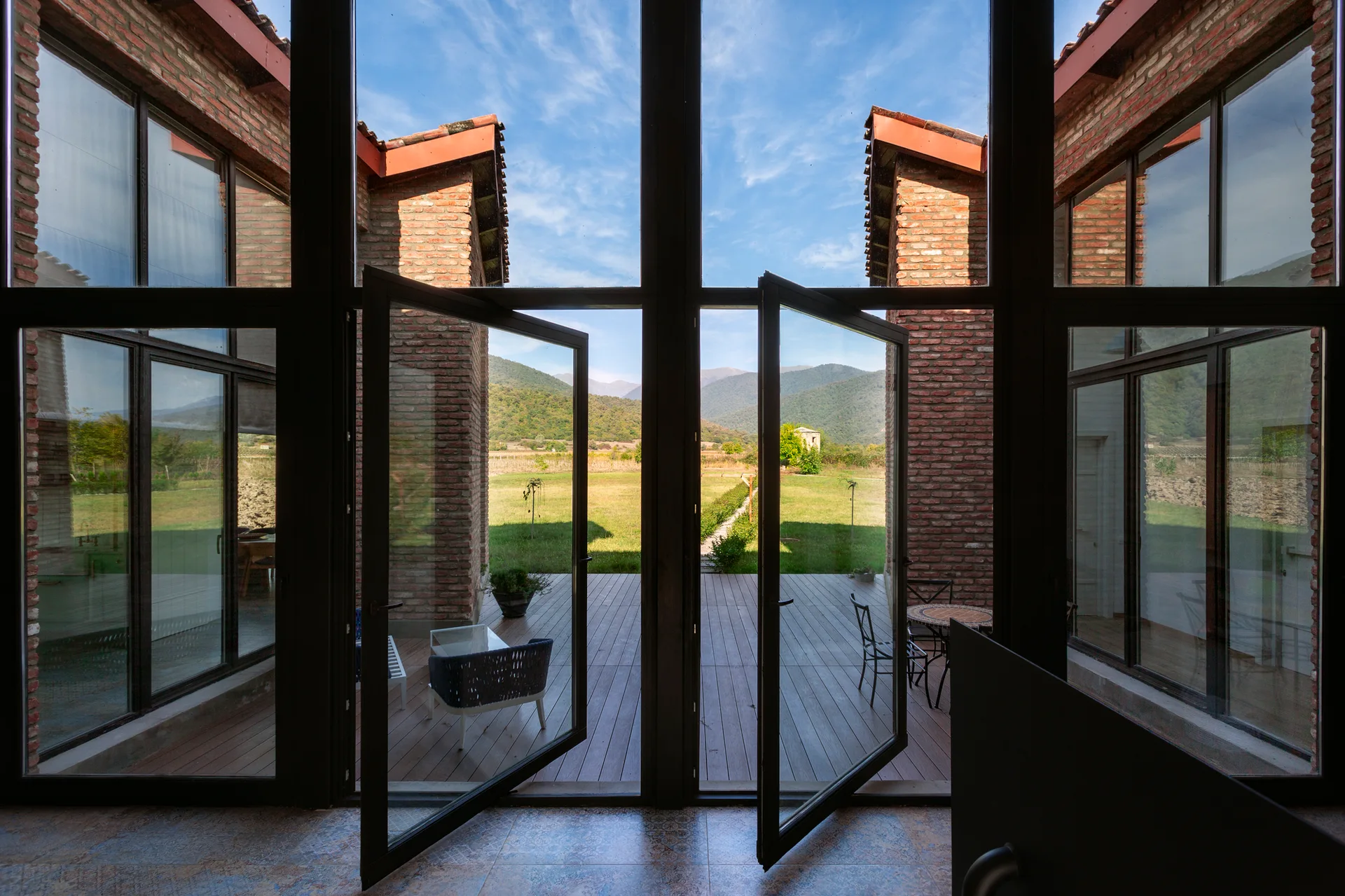 A view through open black-framed glass doors looking out toward a wooden deck with patio furniture, framed by red brick walls under a blue sky.
