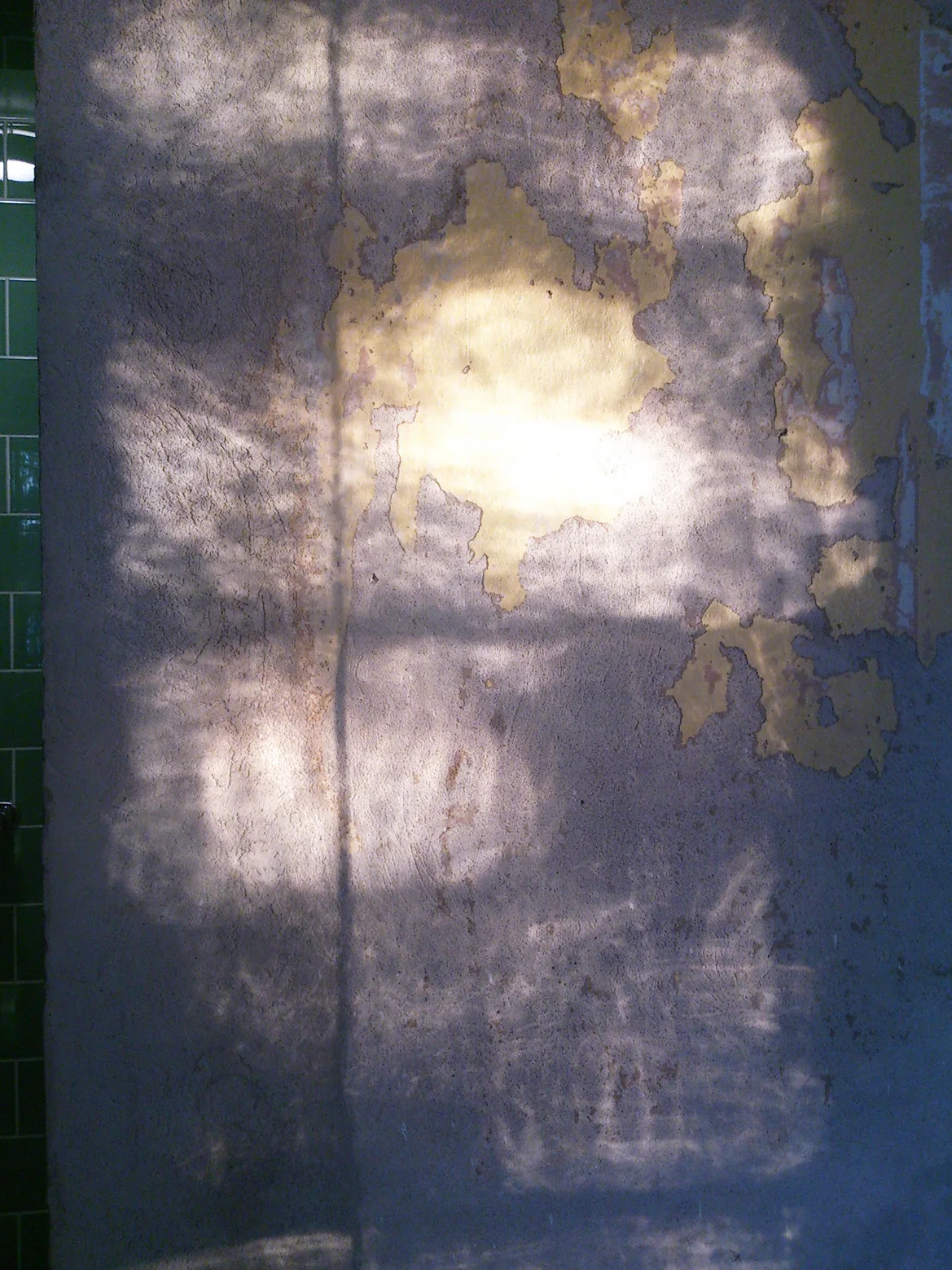 A weathered grey plaster wall with peeling yellow paint features a pattern of light and shadow next to green ceramic wall tiles.