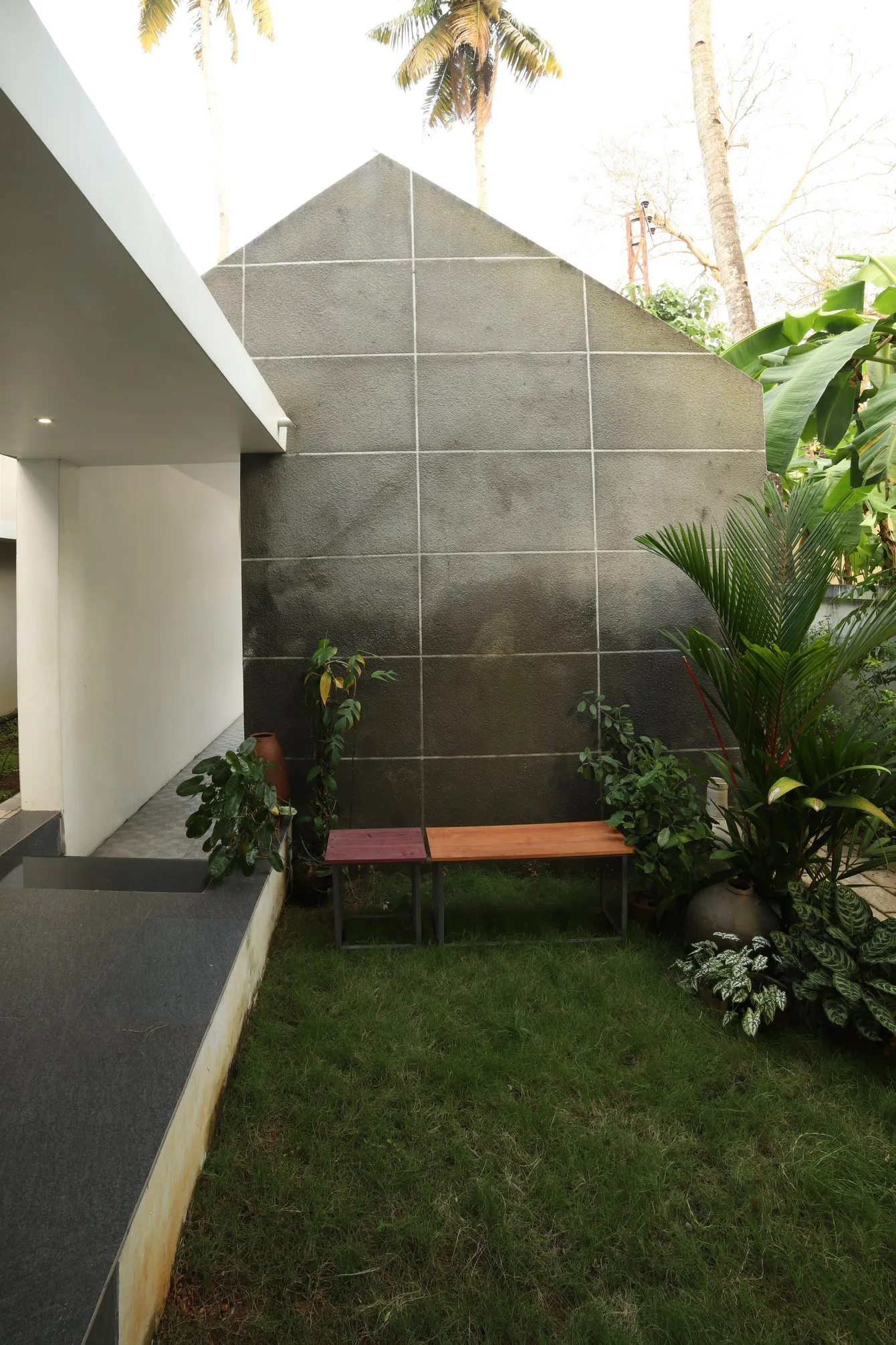 Two wooden benches sit on a small lawn in front of a dark grey stone gabled wall surrounded by lush tropical plants.