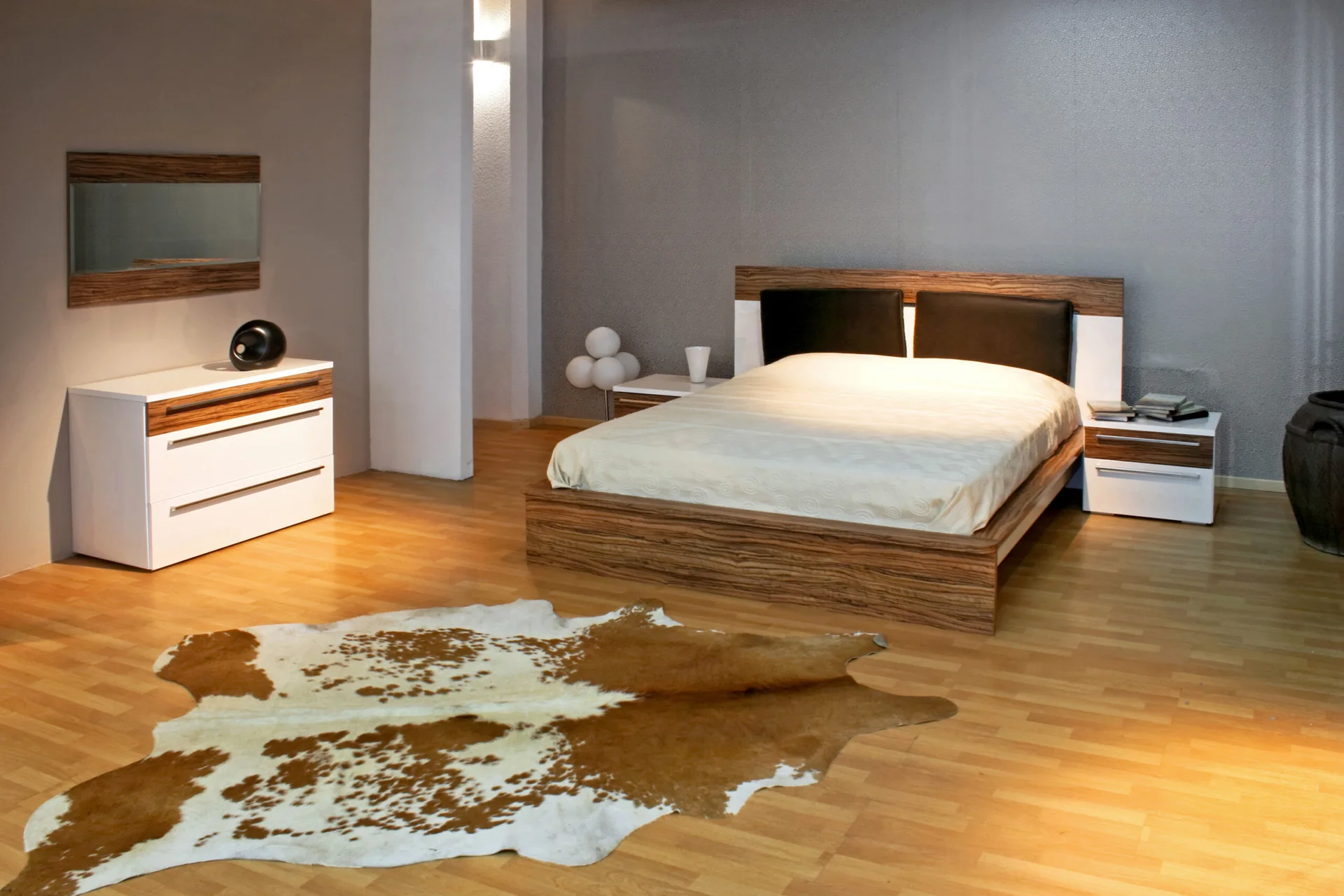 A modern bedroom features a wooden bed frame, matching white and wood nightstands, a matching dresser, and a brown and white cowhide rug.