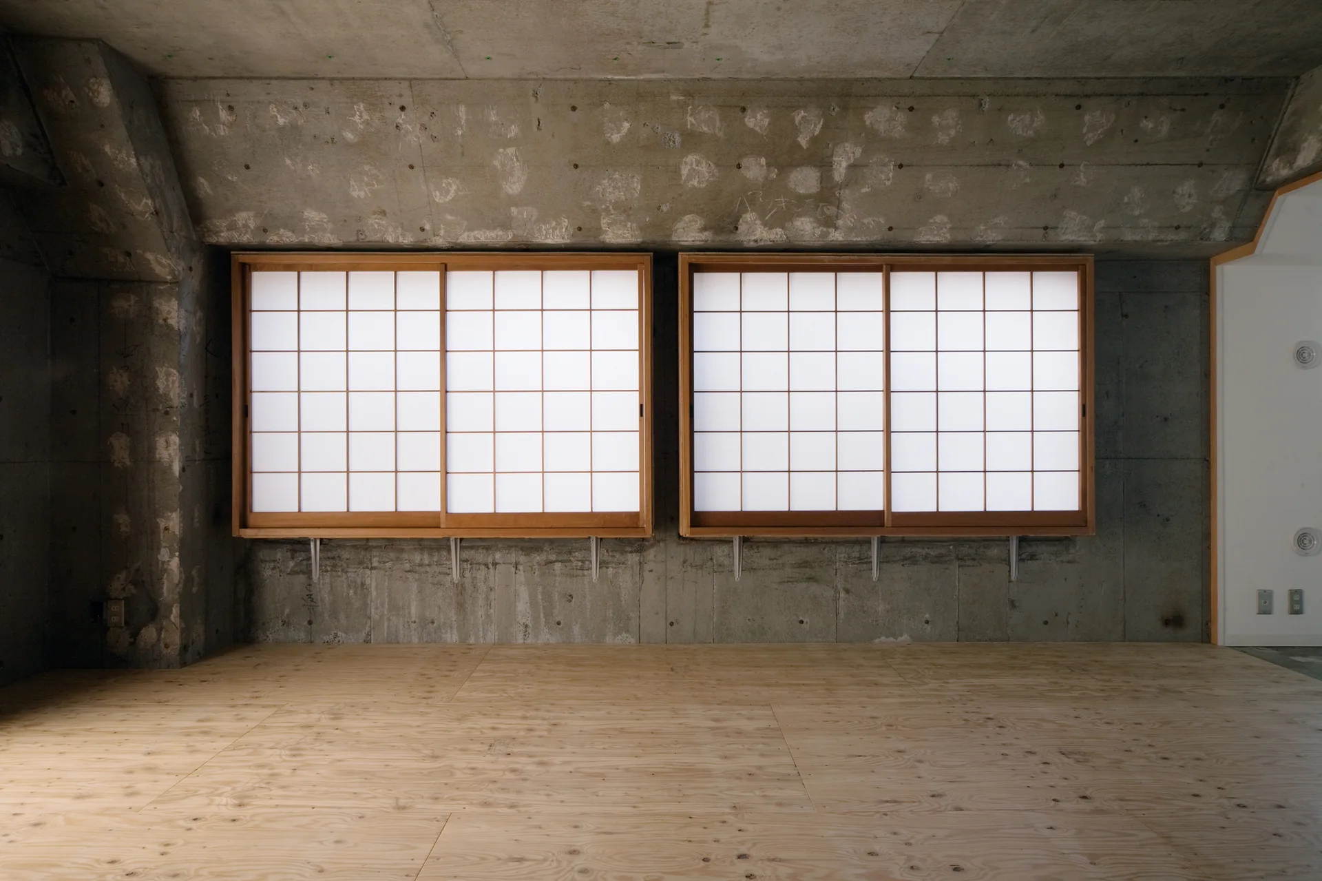 Two wooden shoji sliding windows are mounted against an unfinished concrete wall in an empty room with light plywood flooring.