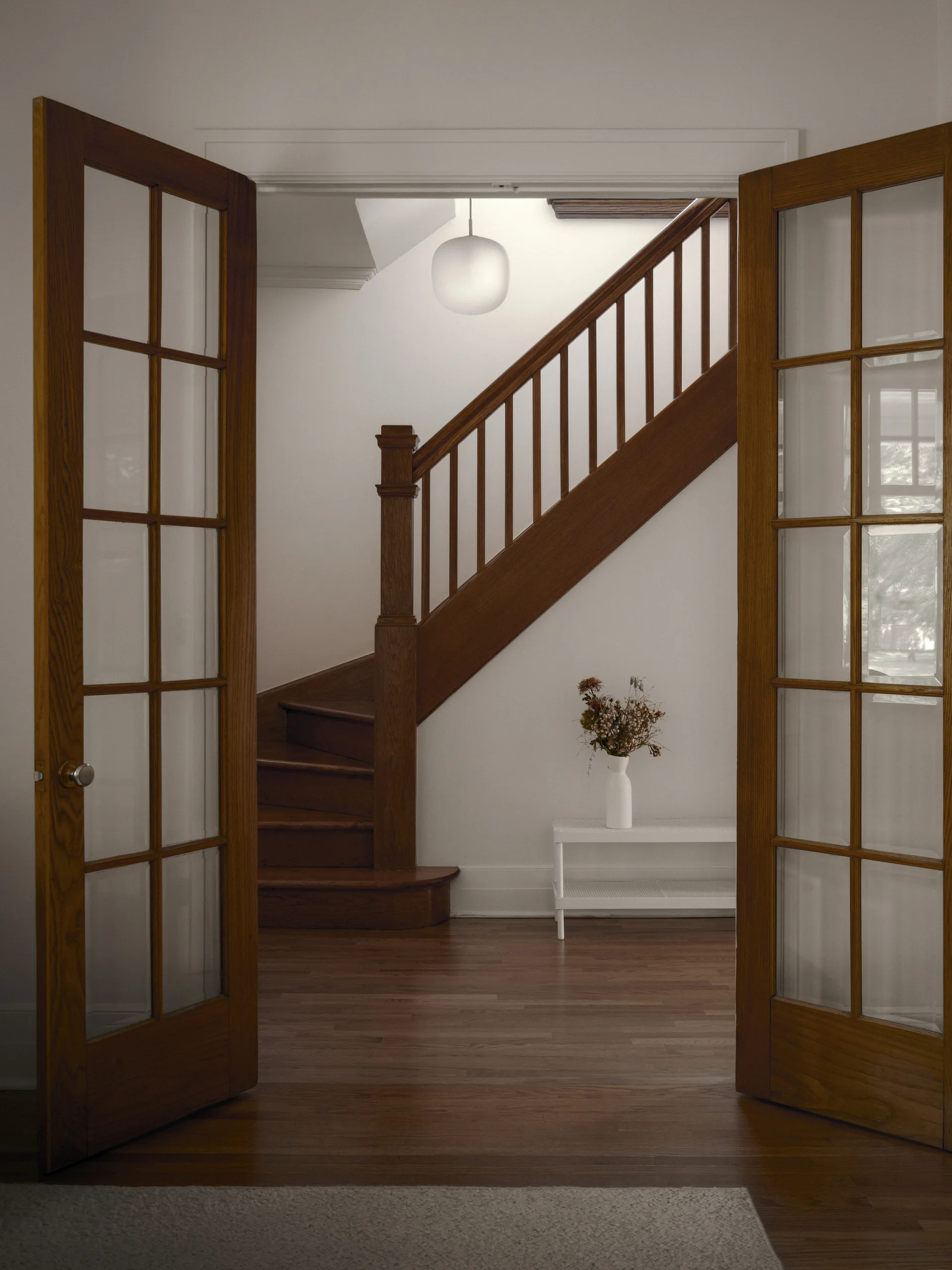 A view through open wood French doors reveals a classic wooden staircase, a white console table with dried flowers, and a globe pendant light.