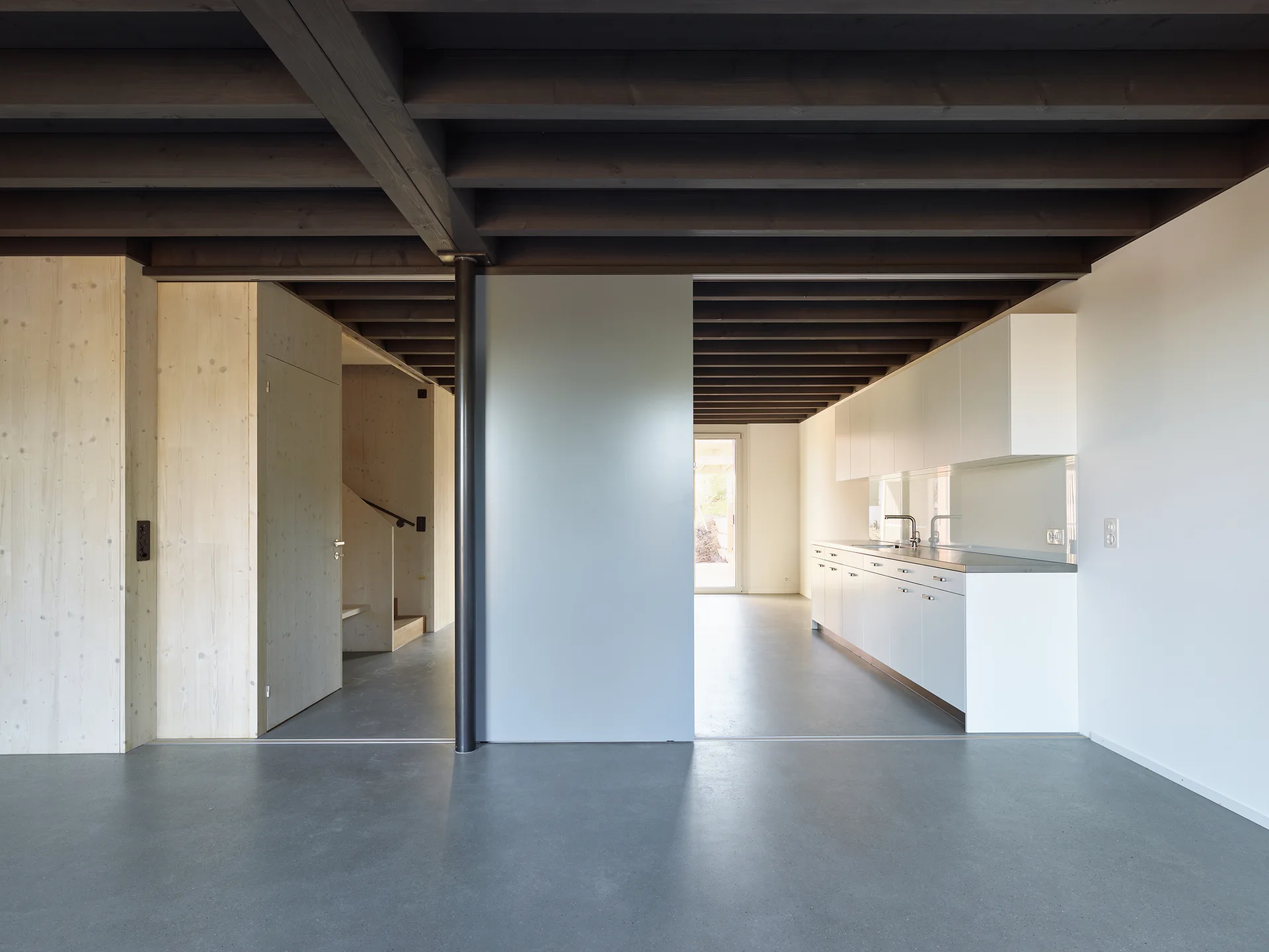 A minimalist kitchen features flat panel white cabinets, a seamless grey concrete floor, and dark exposed wood ceiling beams.
