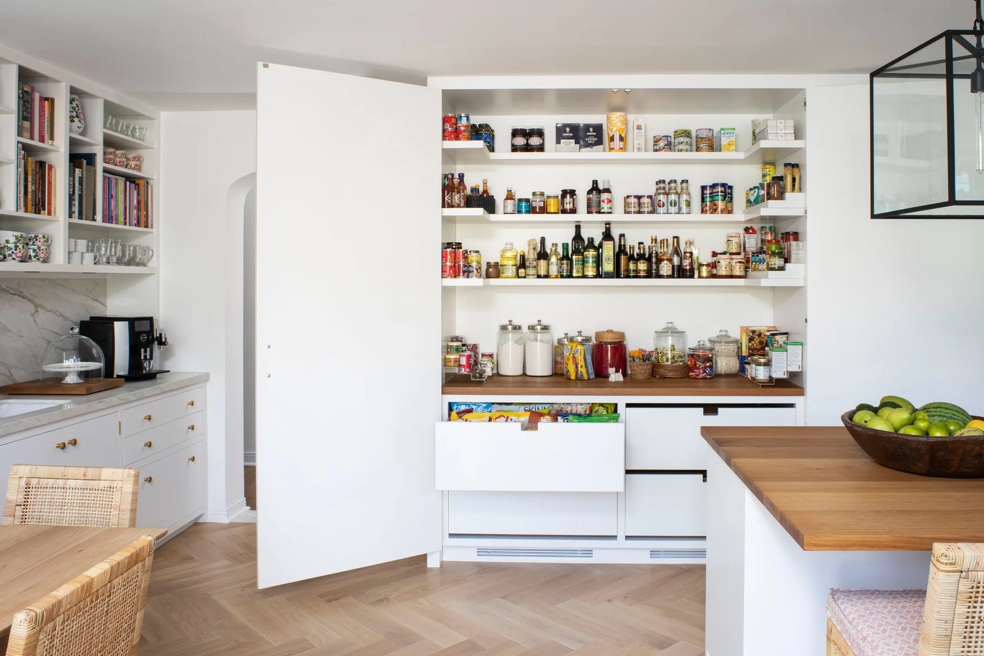 An open white built-in kitchen pantry featuring shelves of jars and cans, a wood countertop, and lower drawers over herringbone wood flooring.