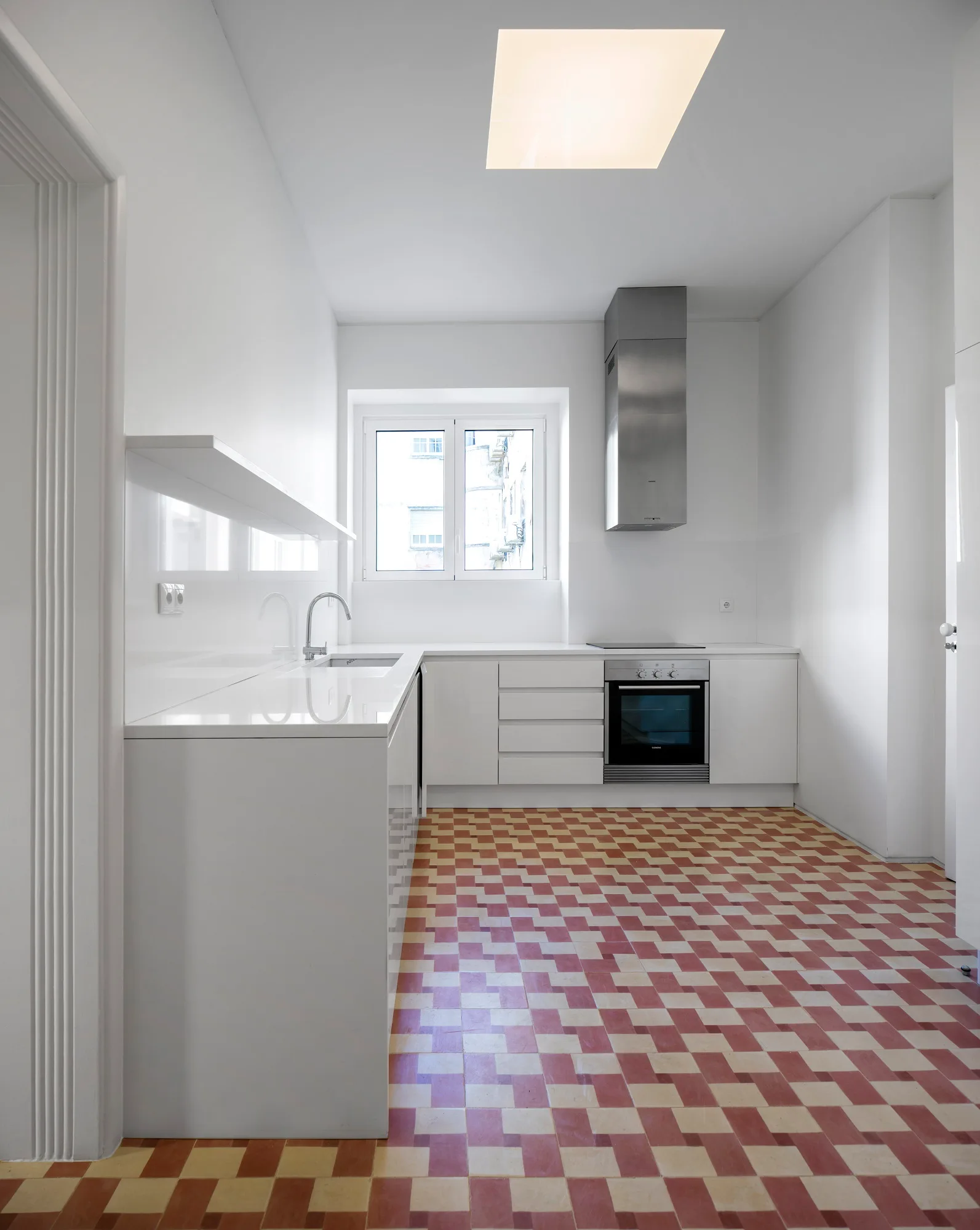 A minimalist white kitchen featuring flat-front cabinets, a stainless steel oven and hood, and a red and cream checkered tile floor.