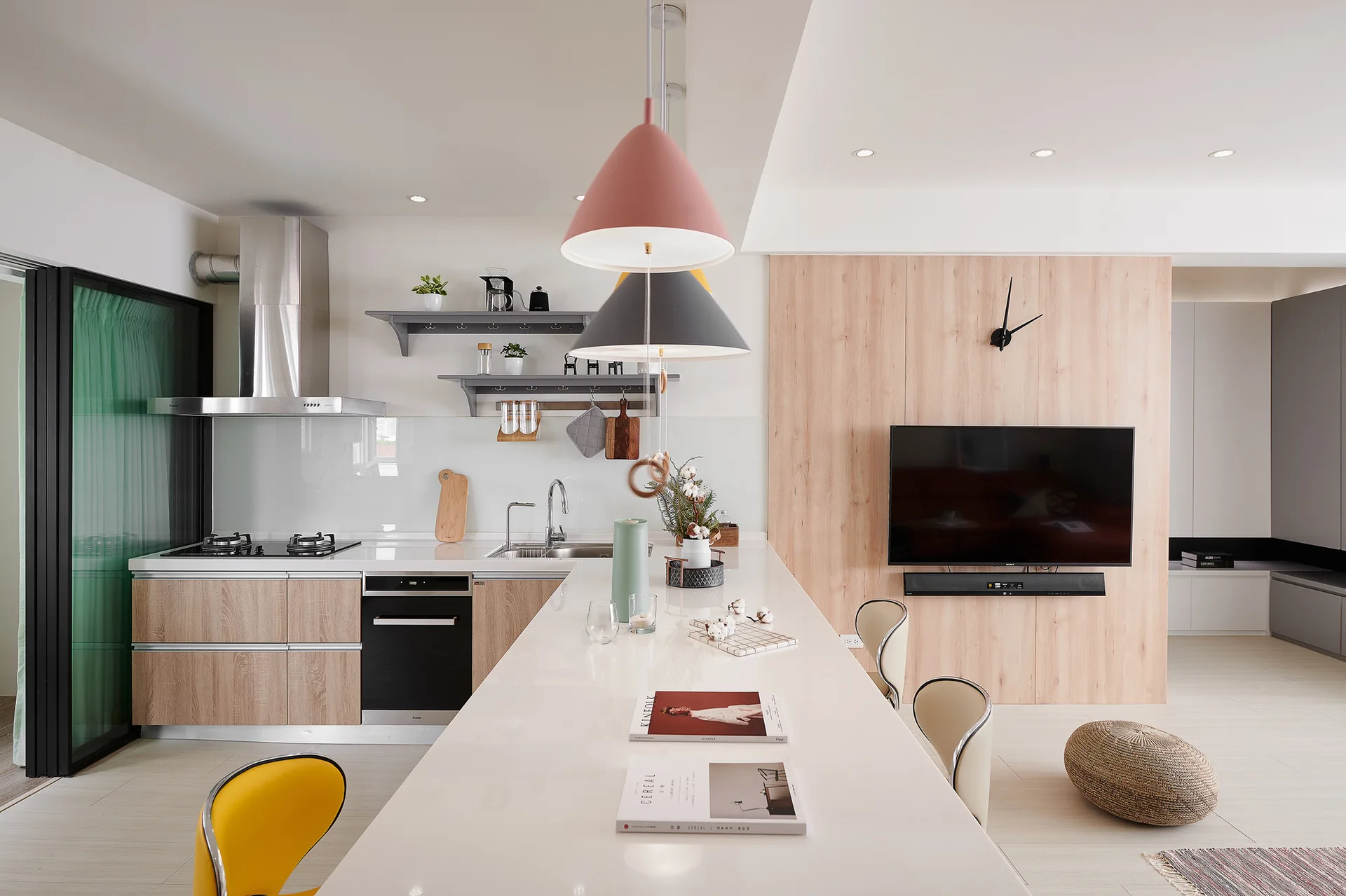 A modern open-concept kitchen features a long white island, light wood cabinetry, and colorful cone pendant lights hanging below a white ceiling.