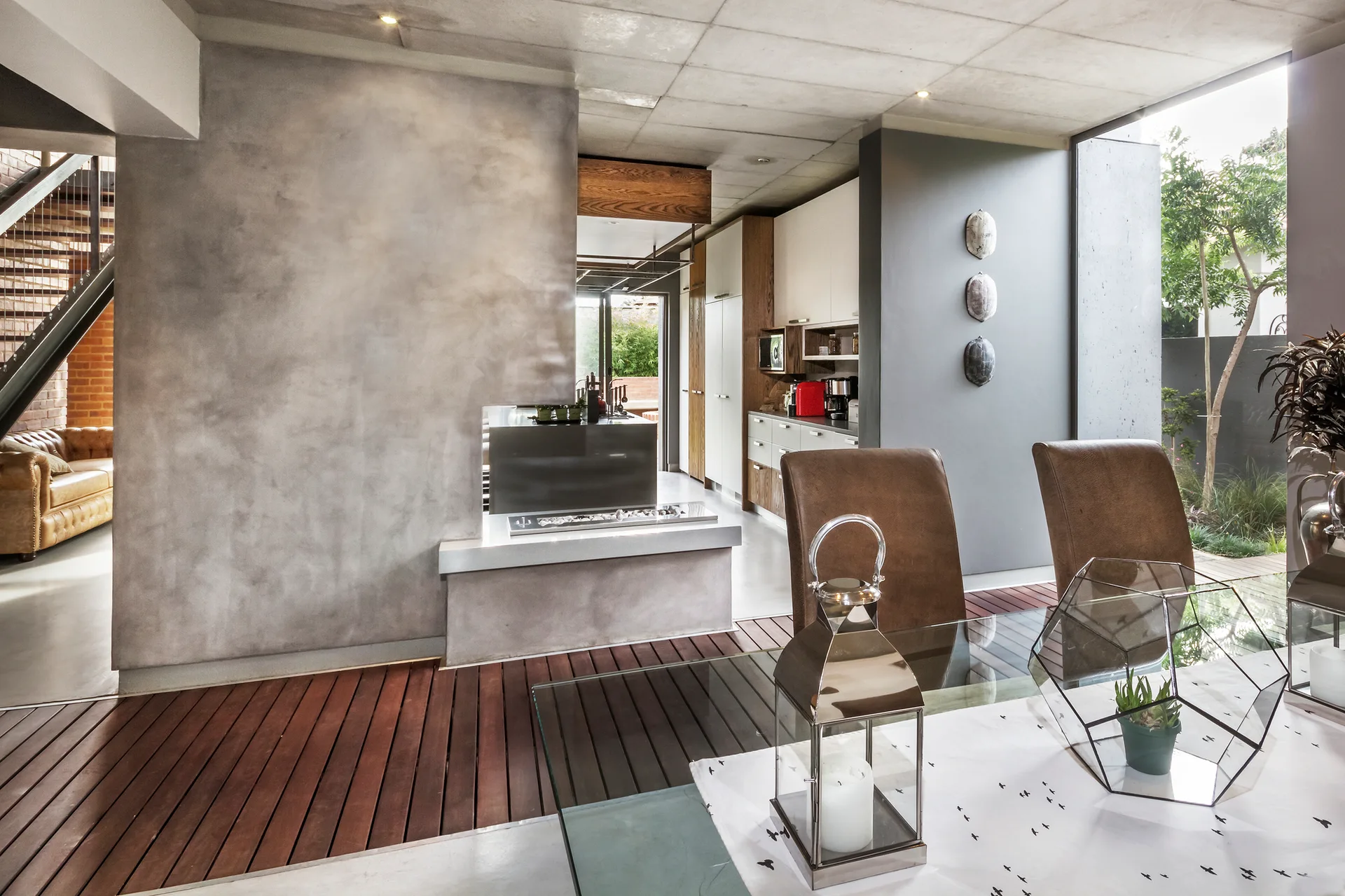 A modern dining room features a glass table with brown leather chairs next to a grey concrete-textured fireplace divider and wood slatted floor inlay.