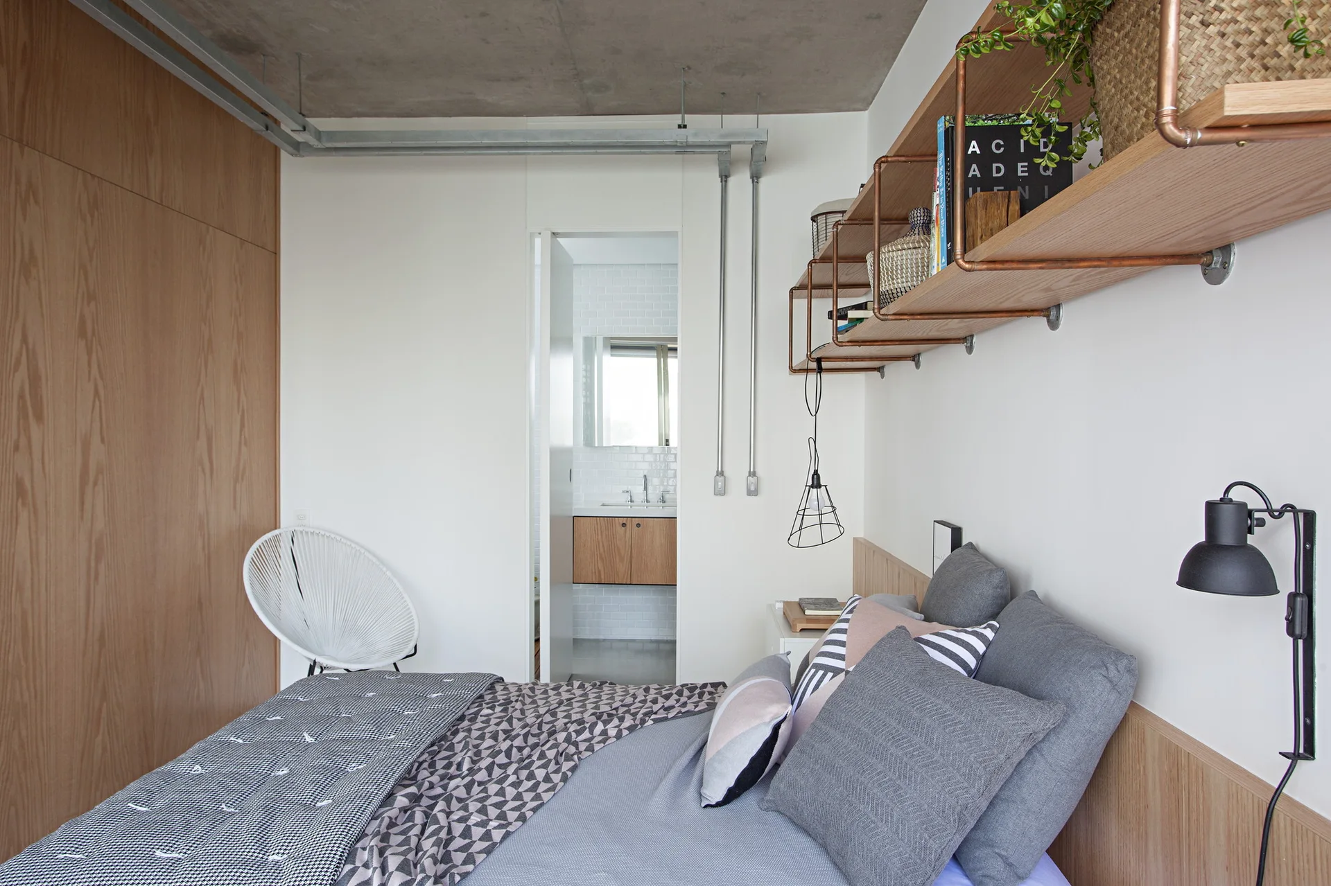 An industrial bedroom with a raw concrete ceiling, copper pipe wall shelves, a wood headboard, and gray bedding.
