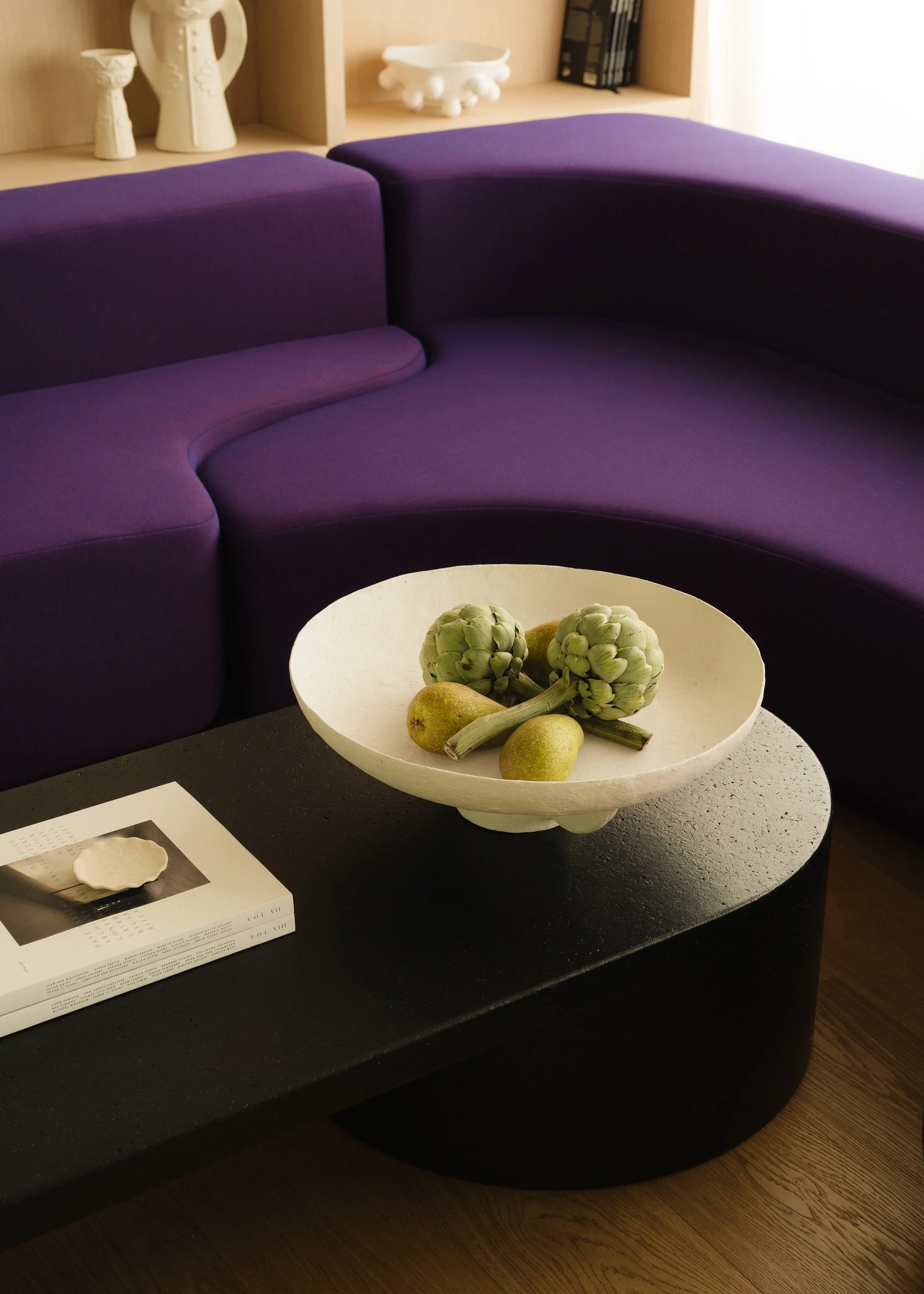 A curved purple sofa stands behind a low black coffee table holding a textured white bowl filled with green artichokes and pears.