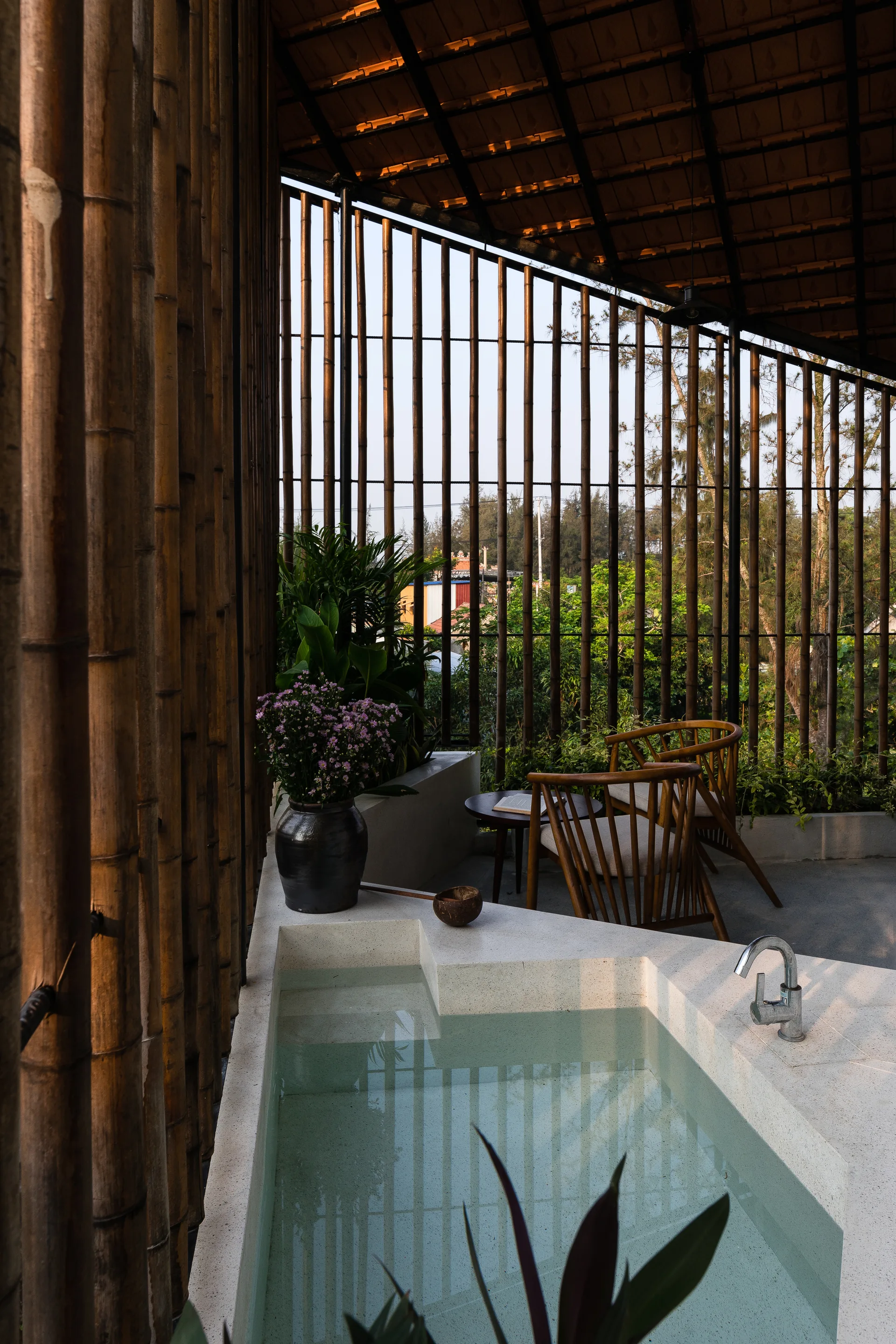 A semi-outdoor patio features a built-in stone bathtub in the foreground, wooden armchairs, and a vertical bamboo screen wall.